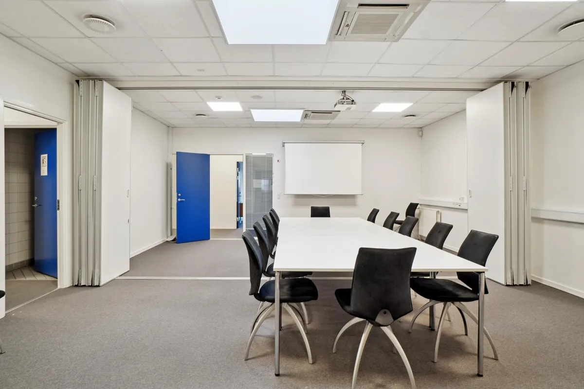 Langt konferencebord med flere stole omkring, hvidt bord og mørke stole, lyst hvidt loft, hvidt vægmaleri og projektor foran