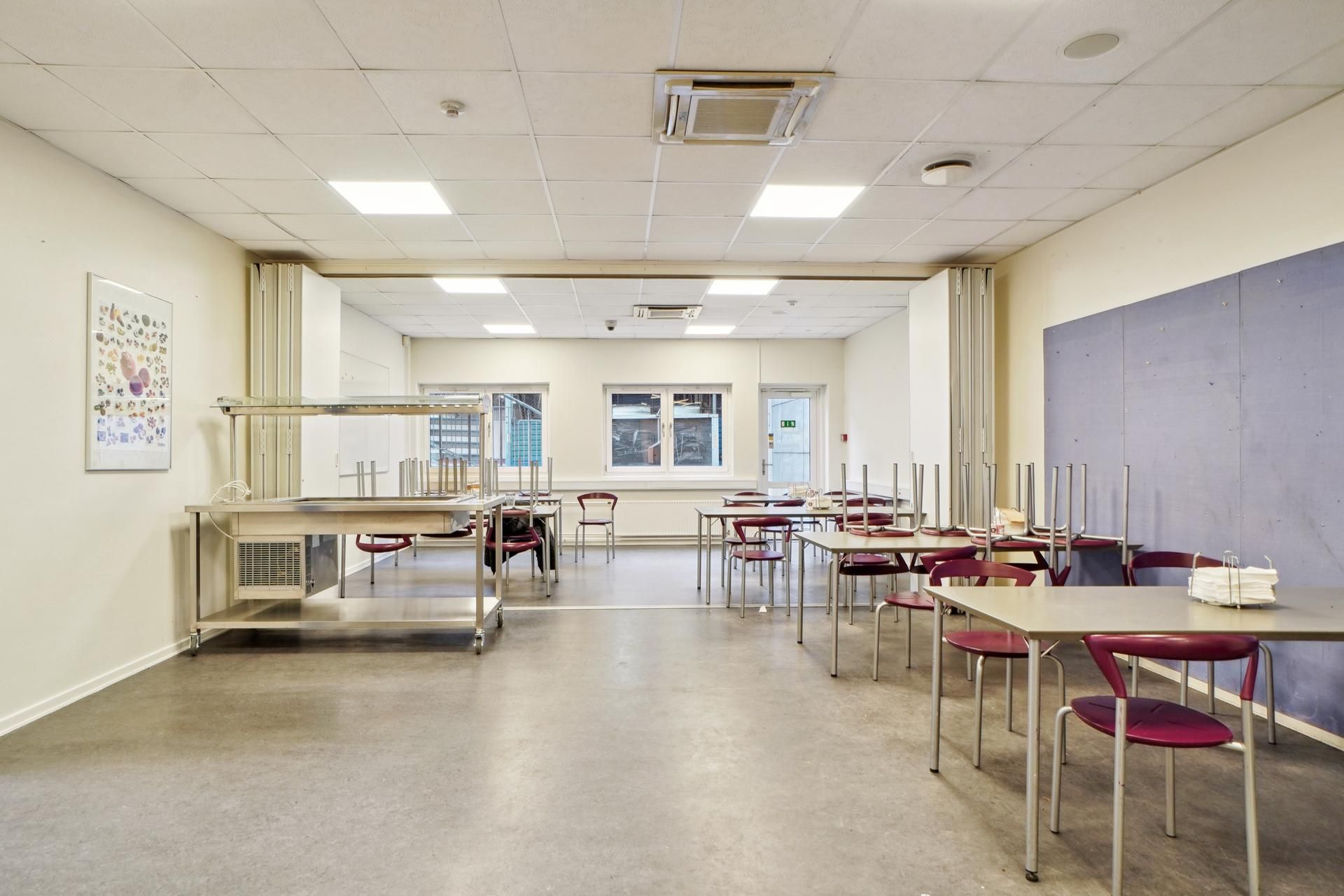 Indeni er der et tomt lokale med flere borde og stole samt et bord med høj hylde og et billede på væggen