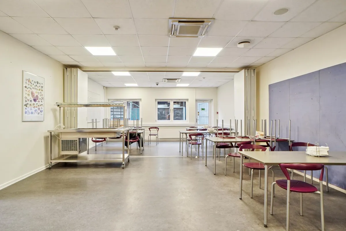 Indeni er der et tomt lokale med flere borde og stole samt et bord med høj hylde og et billede på væggen