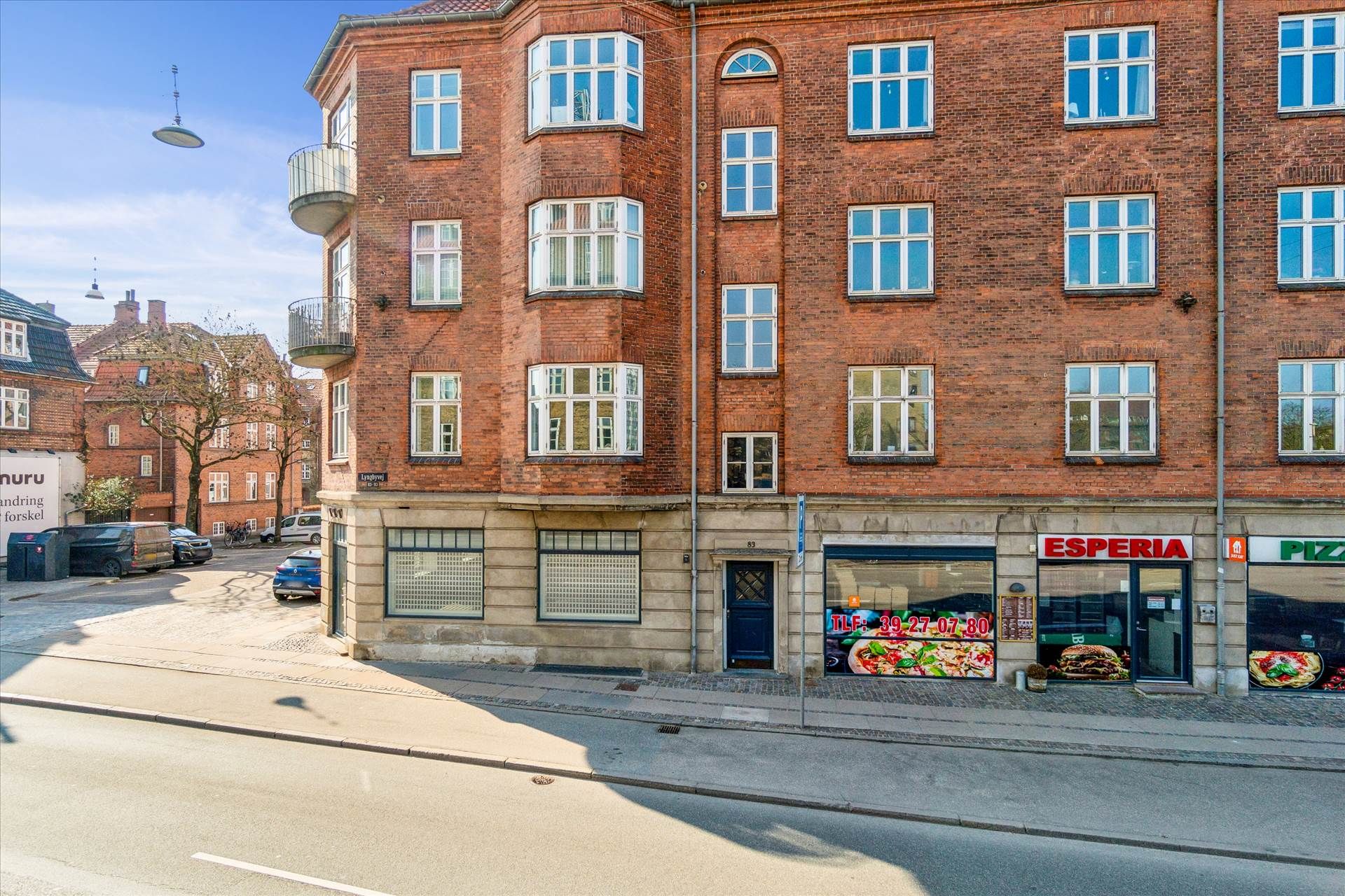 En to-etagers lejlighedsbygning med røde mursten og mange vinduer og balkoner, samt en restaurant og pizzeria med en pizza