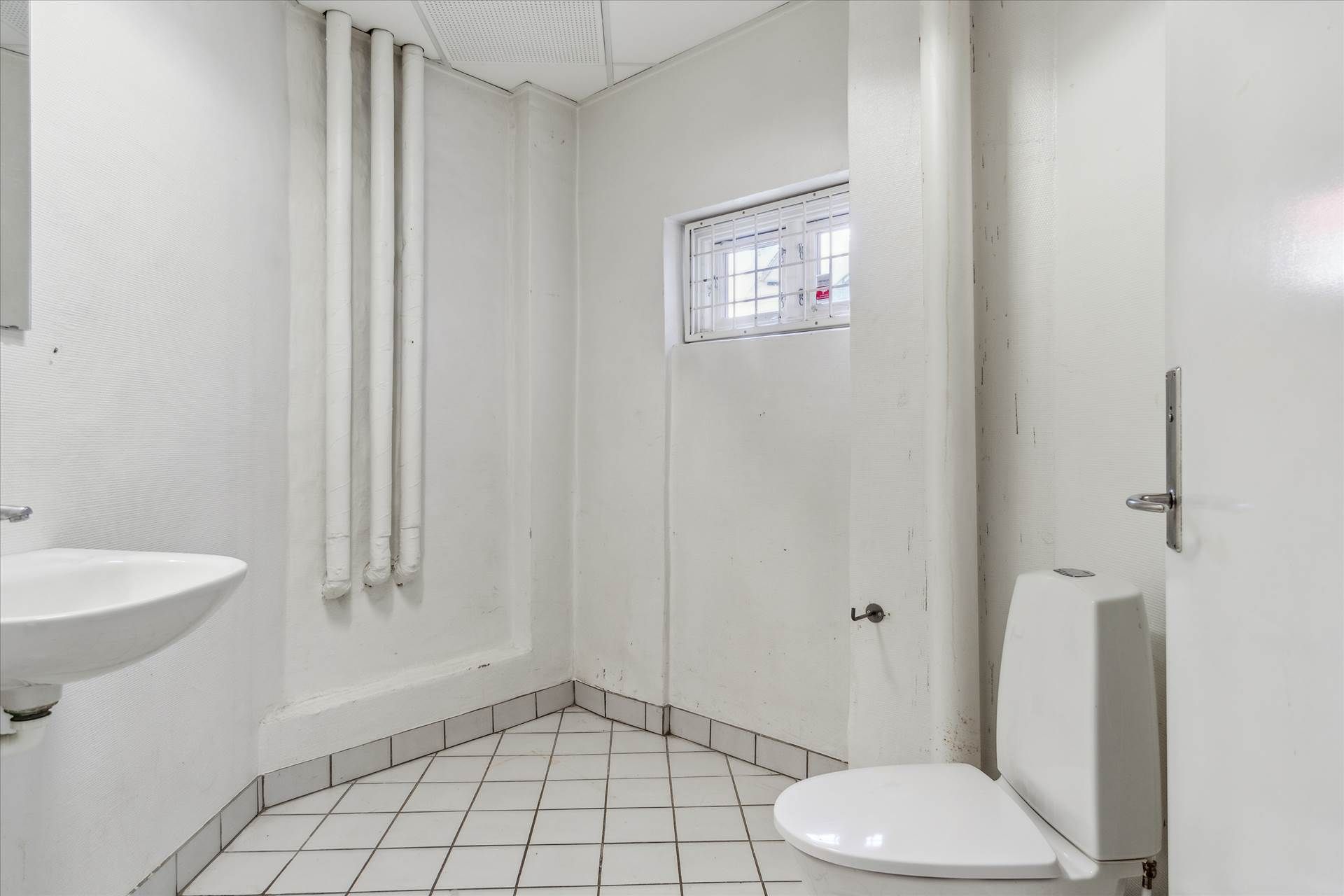 En lille hvid toilet med toiletbøjle og et hjørnevindue samt en hvid vask med en håndvask.