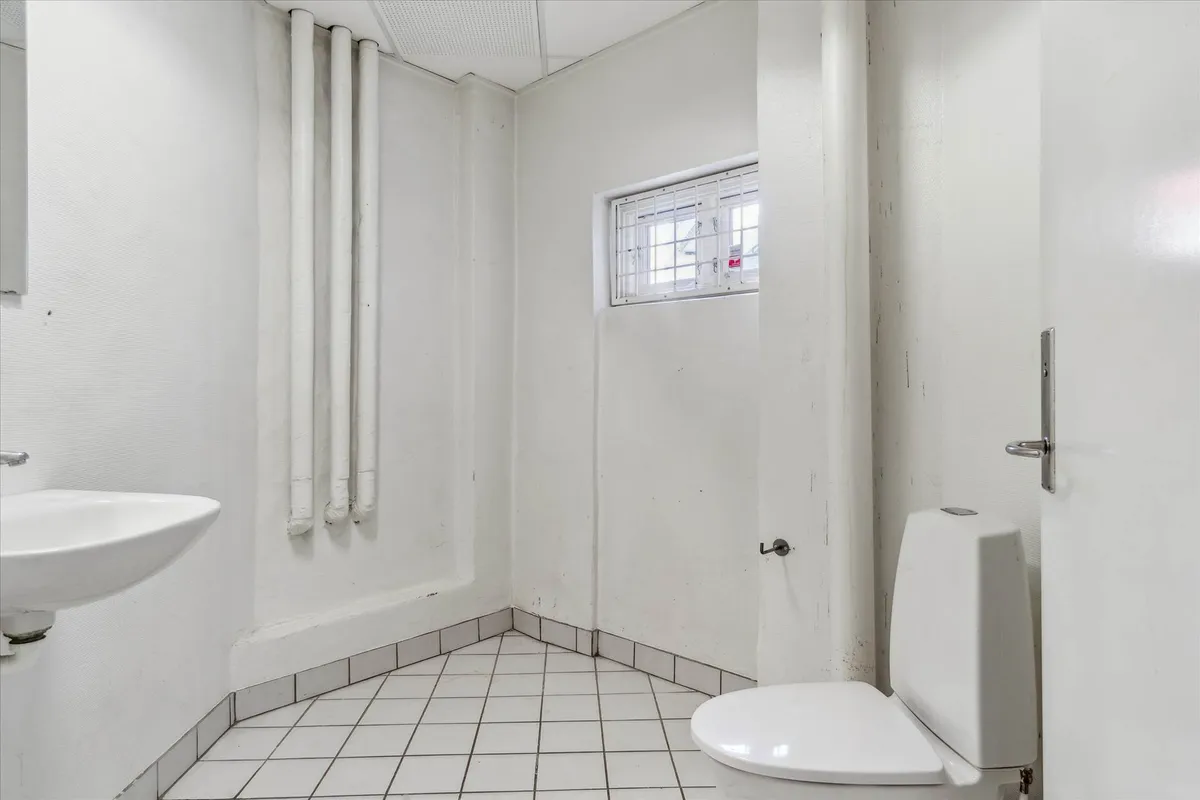 En lille hvid toilet med toiletbøjle og et hjørnevindue samt en hvid vask med en håndvask.