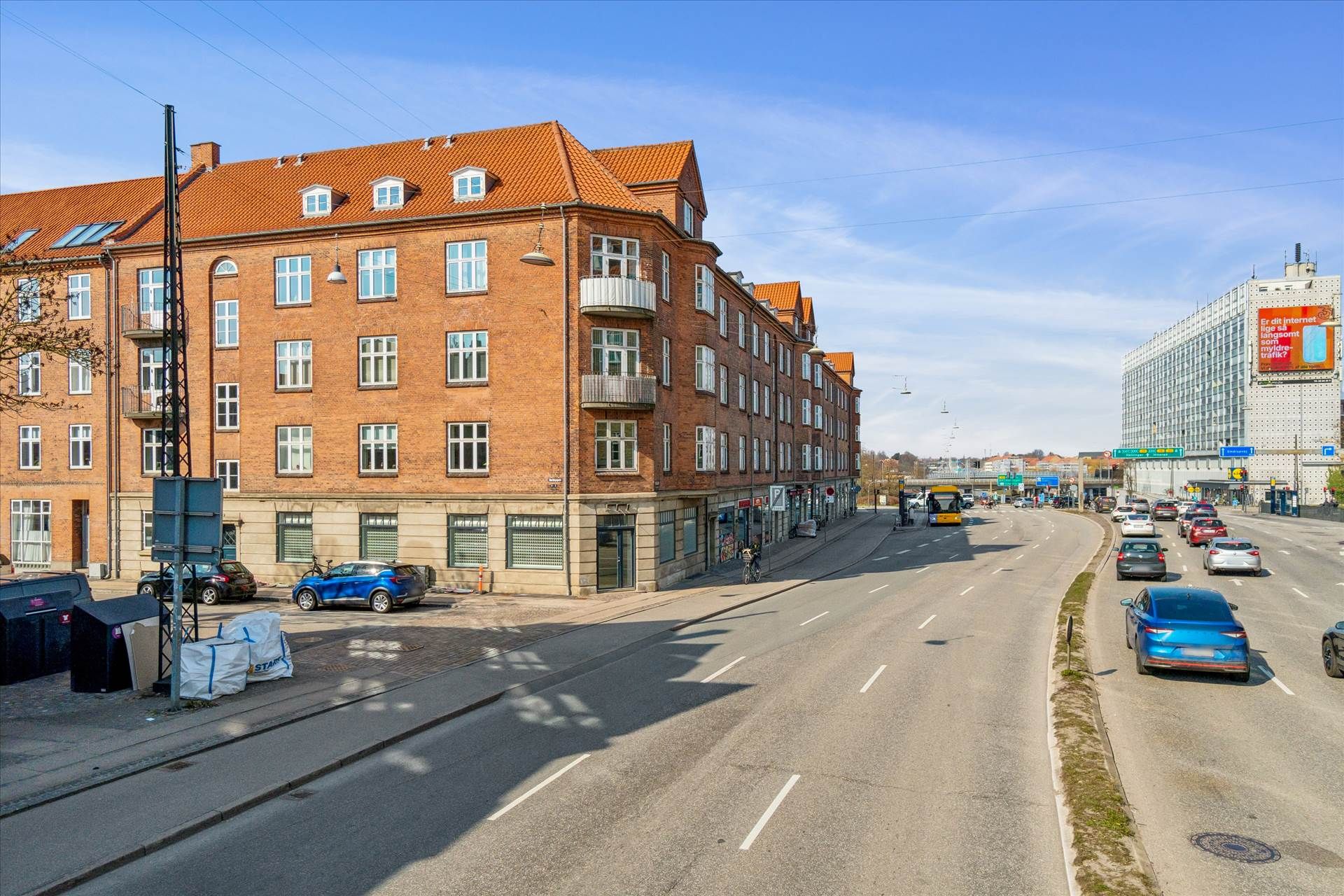 Butik og parkeringsplads på hjørnet af Lyngbyvej og Bortigsgade i København med biler og busser på vejen og et skilt med