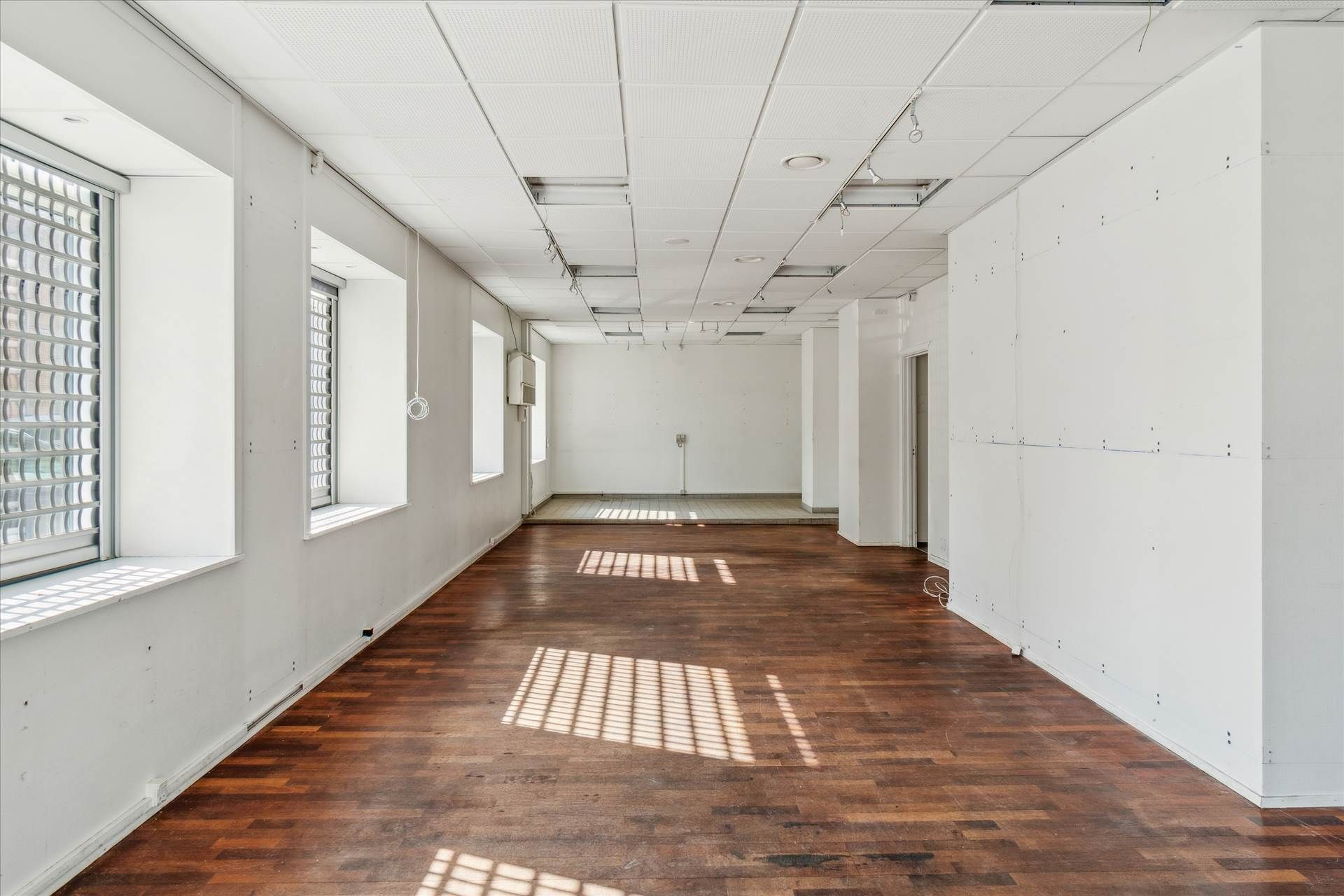 En stor og tom kontorejendom med hårdt trægulv, hvide vægge og vinduer med skyggekast indrammet af hvide ruder