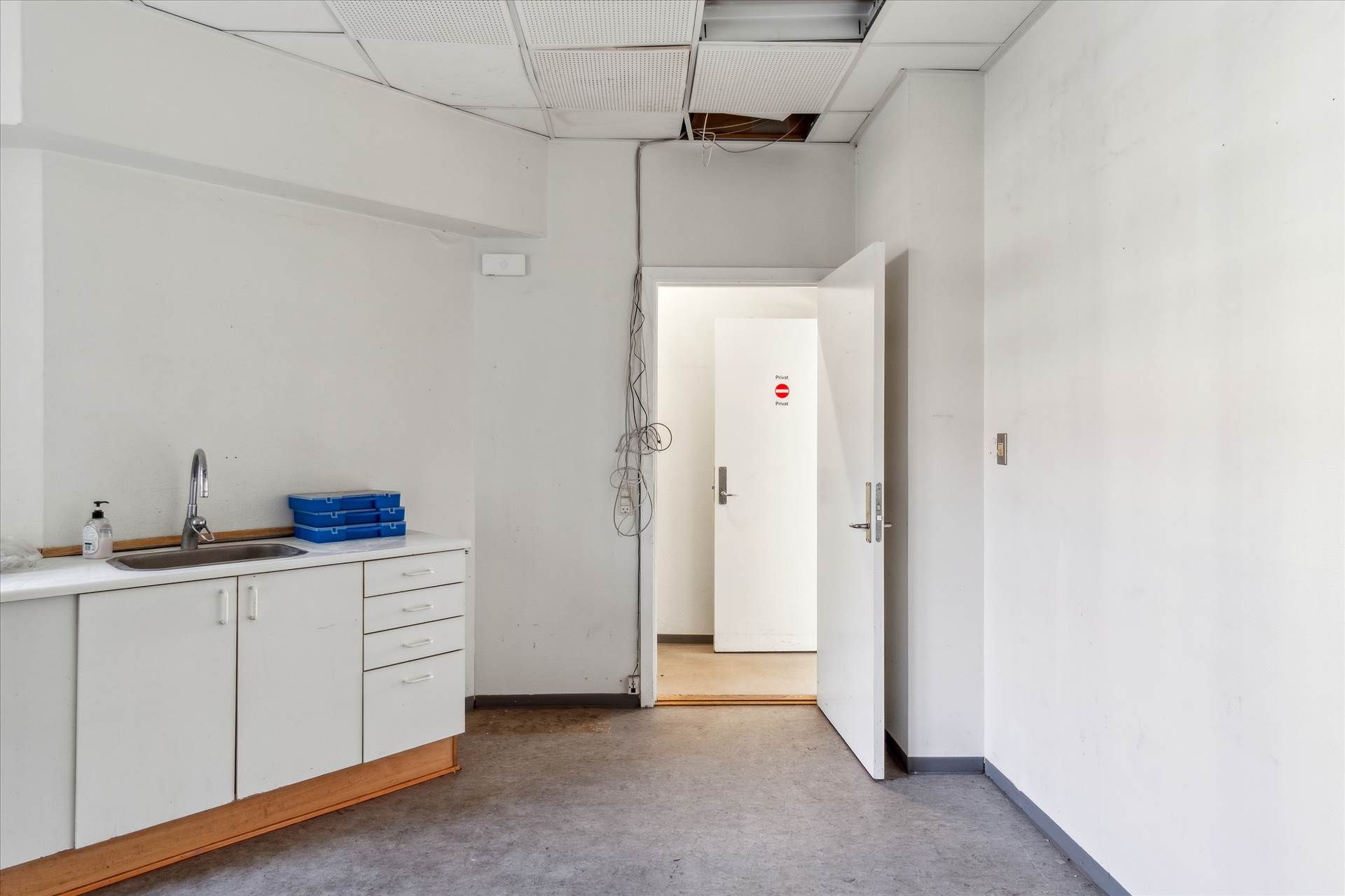 Tomt rum med hvidt loft og vægge, åben dør, sink med vandhane og sæbeflaske, og rødt skilt med teksten Privat
