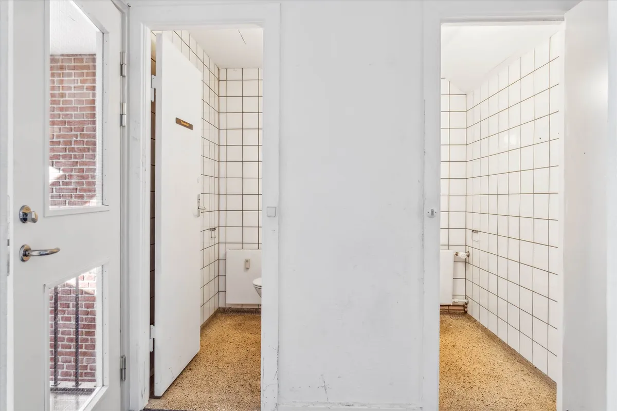 To toiletrum med hvide murfliser og gulv samt toilet og hængetoilet i hvert rum