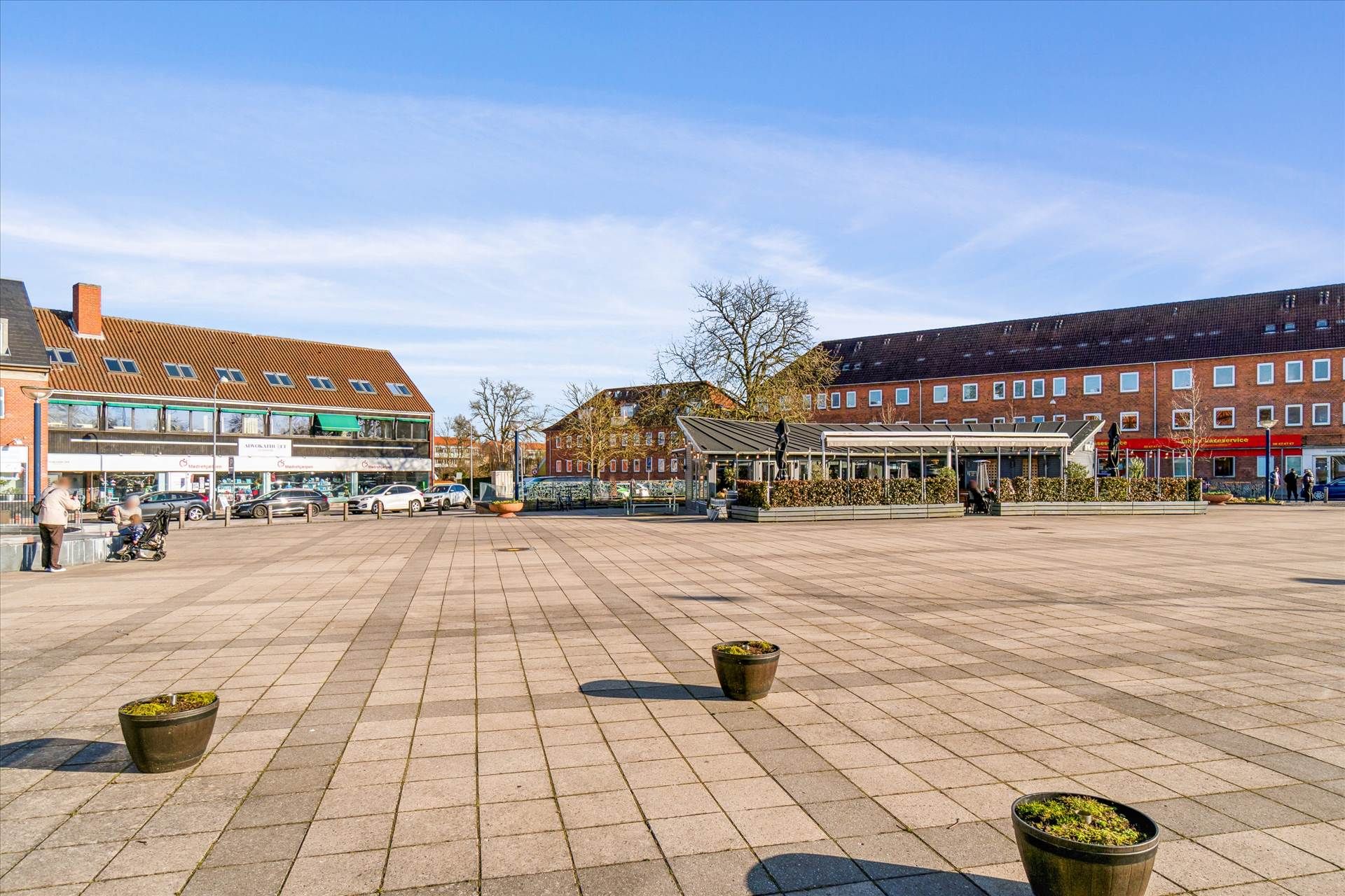 En stor torv med butikker og caféer, hvor to personer står med en barnevogn, og to kvinder går forbi en bil.