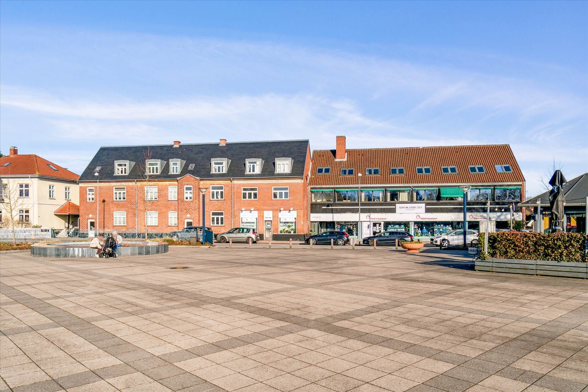 En åben gågade med butikker og caféer. Foran er en familie med barnevogn og en stor parkeringsplads med biler og planter.