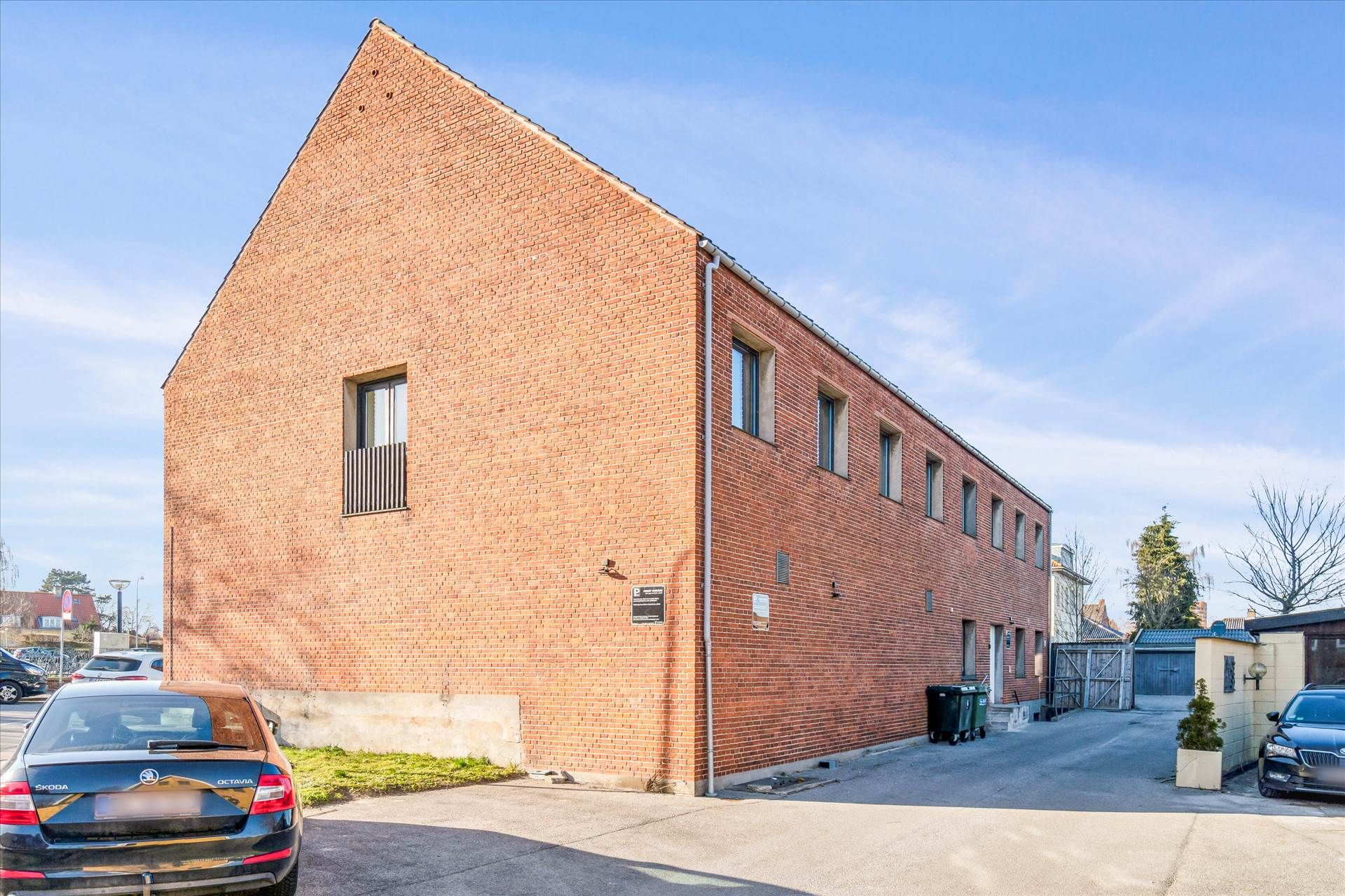Kontor med røde mursten og glasvinduer, med en bilparkeringsplads og grønne skraldespande.
