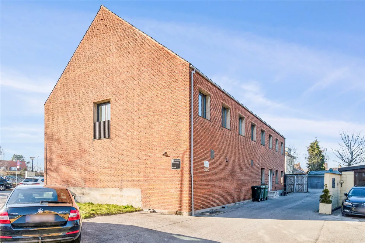 Kontor med røde mursten og glasvinduer, med en bilparkeringsplads og grønne skraldespande.