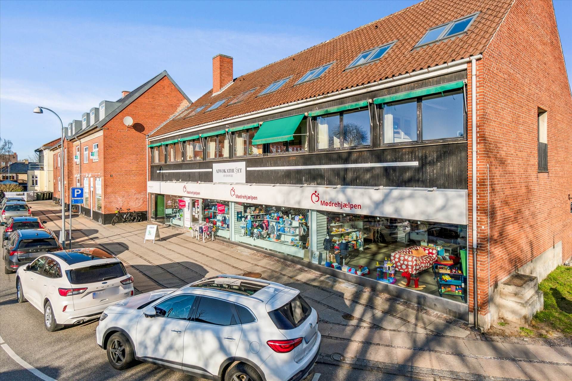 Flere hvide SUV'er parkeret på en parkeringsplads, udsigt over en butik med vinduer, skilte og skrifttegn, solskin og skyer.