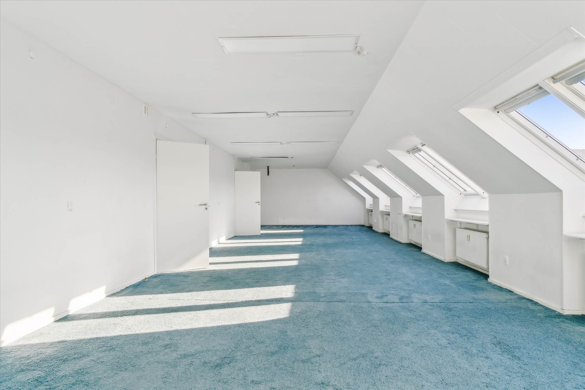 Rummelig loftsværelse med blå gulvtæppe og hvide vægge, samt loftsvinduer og hvide døre, der stiger op mod loftet.