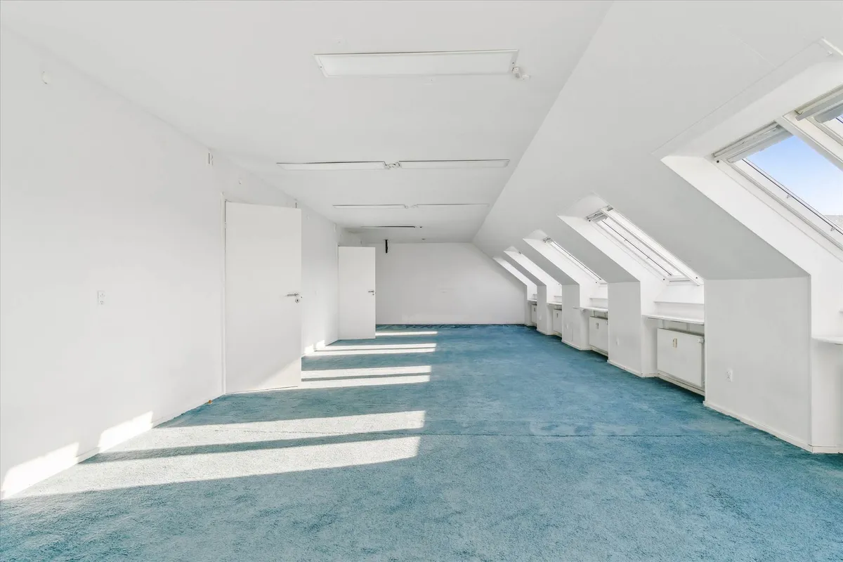 Rummelig loftsværelse med blå gulvtæppe og hvide vægge, samt loftsvinduer og hvide døre, der stiger op mod loftet.
