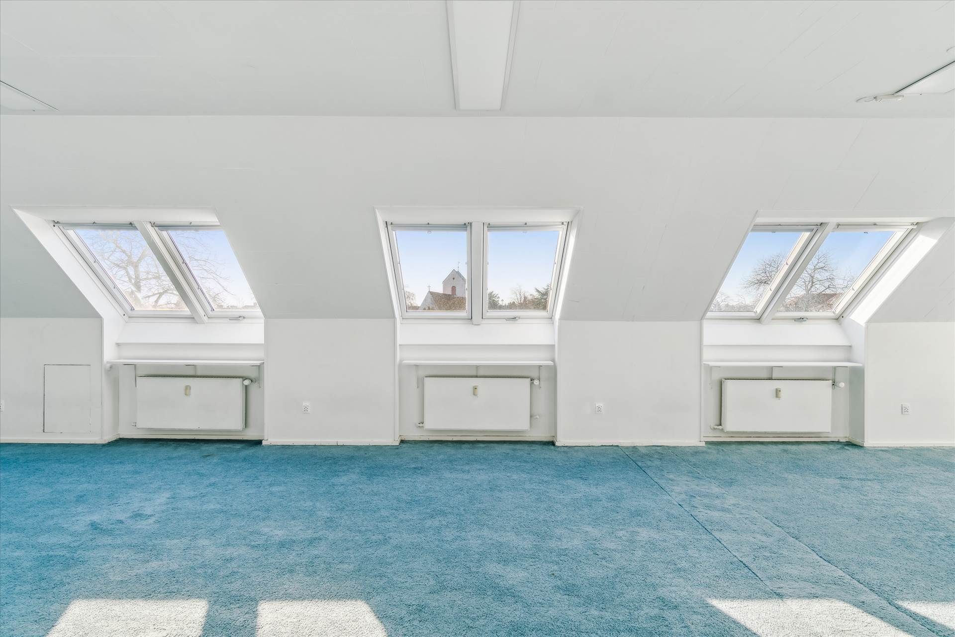 Et lyst og rummeligt værelse med lysegul gulvbelægning og tre store loftsvinduer. Der er hvide vægge og hvide radiatorer