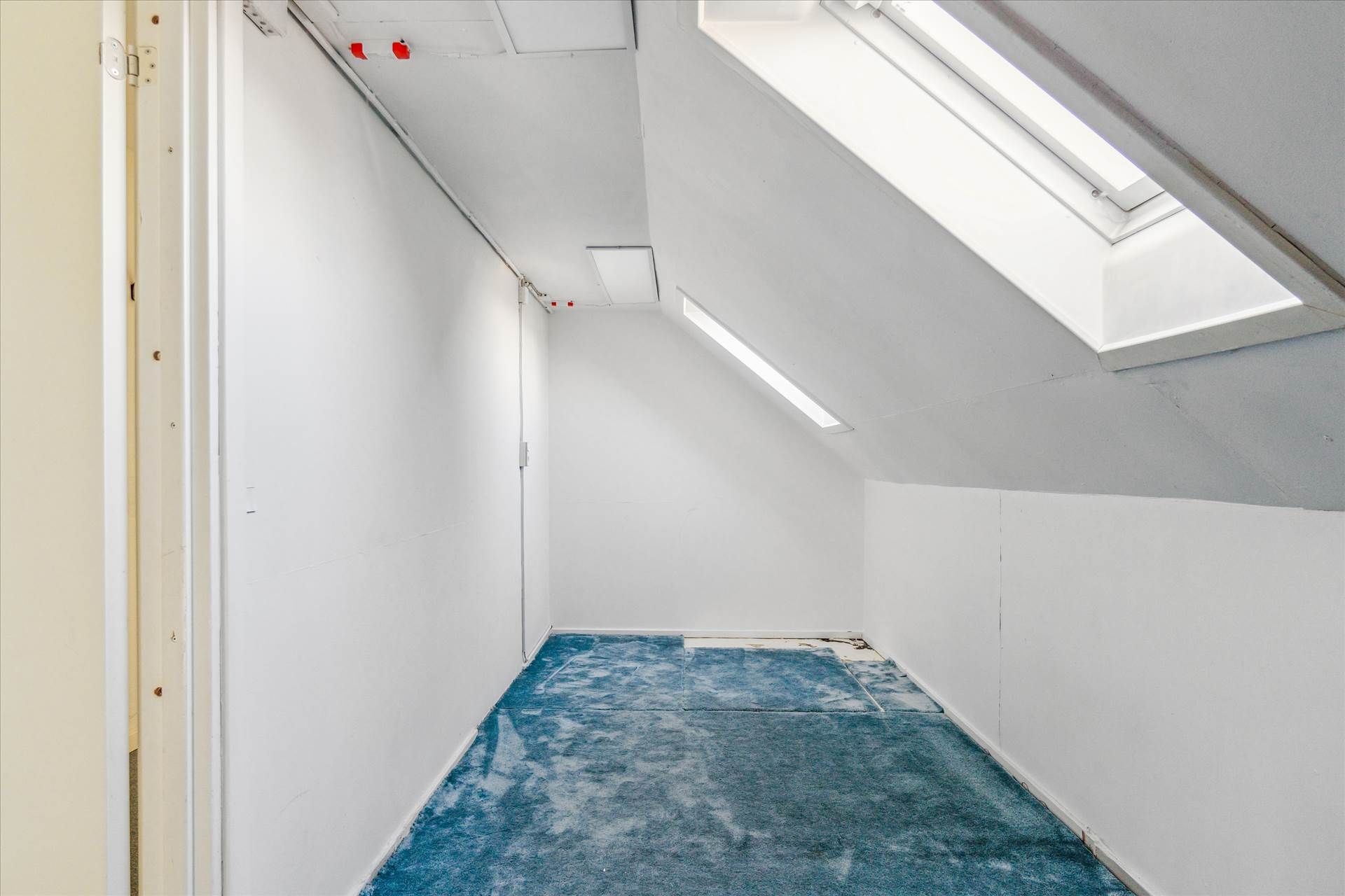 En stor, tom rum med lyse lofter, hvide vægge og blå gulvbelægning. Der er to loftsvinduer og en dør, der er delvis åben.