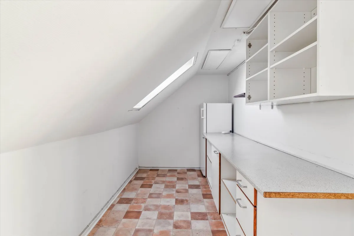 Hjørne loftsværelse med køkkendel, hvidt loft og vægge, loftsvindue og hvidt køleskab med møbler og bordplade