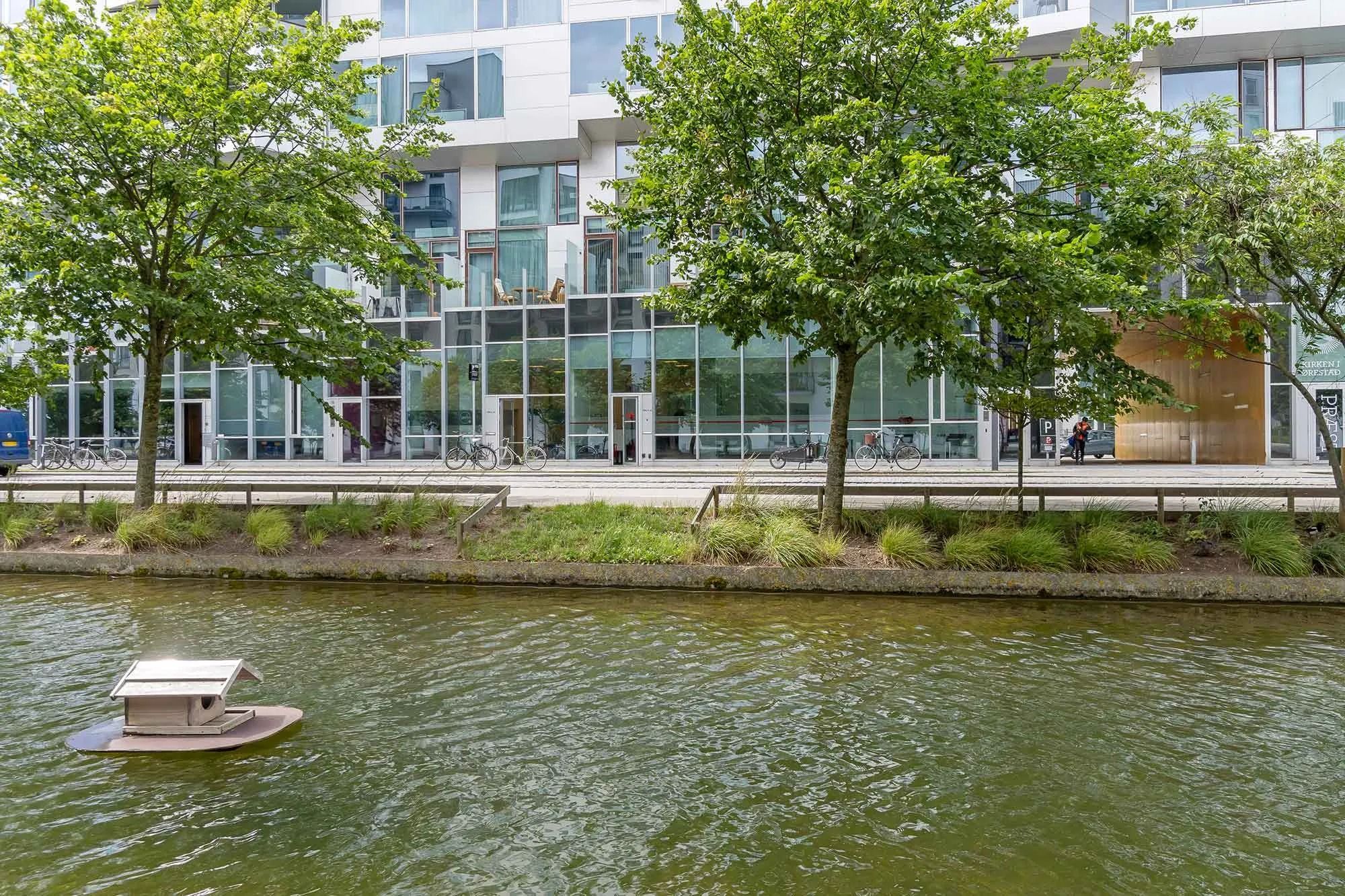 En smuk ejendom med glasvinduer og træer med vandløb i Amsterdam. Et lille træhus er flydende på vandet og en person går