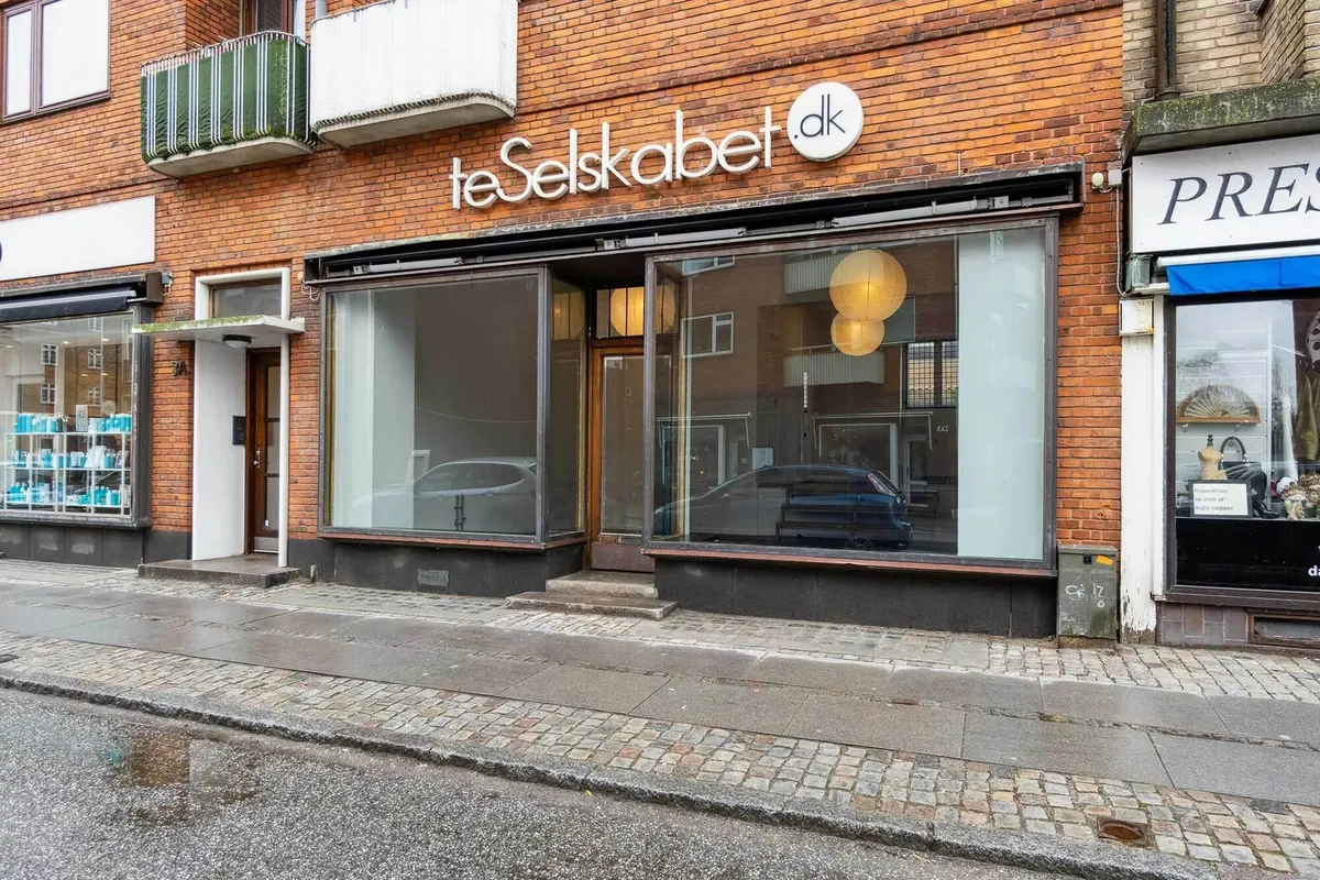 Lejlighedsbygning med store vinduer og butikker, hvor den ene butik hedder teSelskabet.dk og den anden hedder Press. Der er