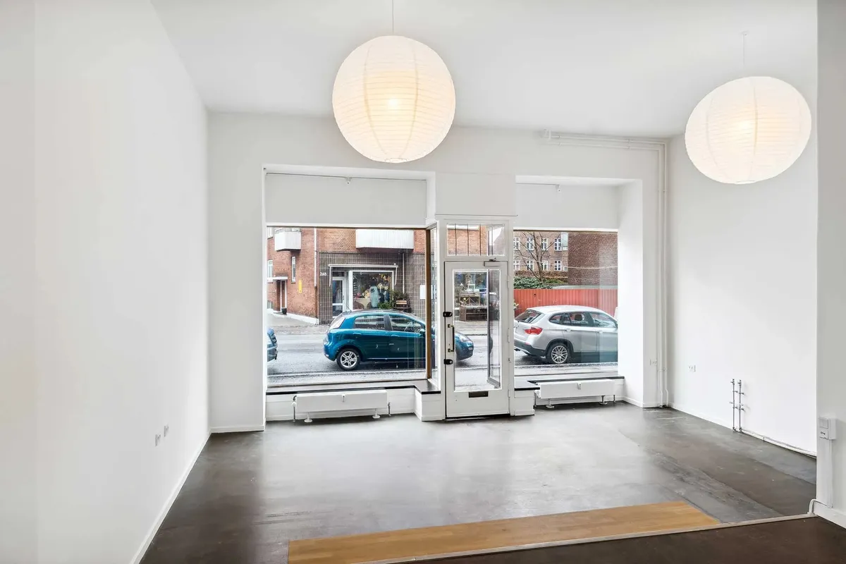 En tom, hvid malet butik med store vinduer og hvide rundede loftslampe. To biler parkeret udenfor og røde huse i baggrunden