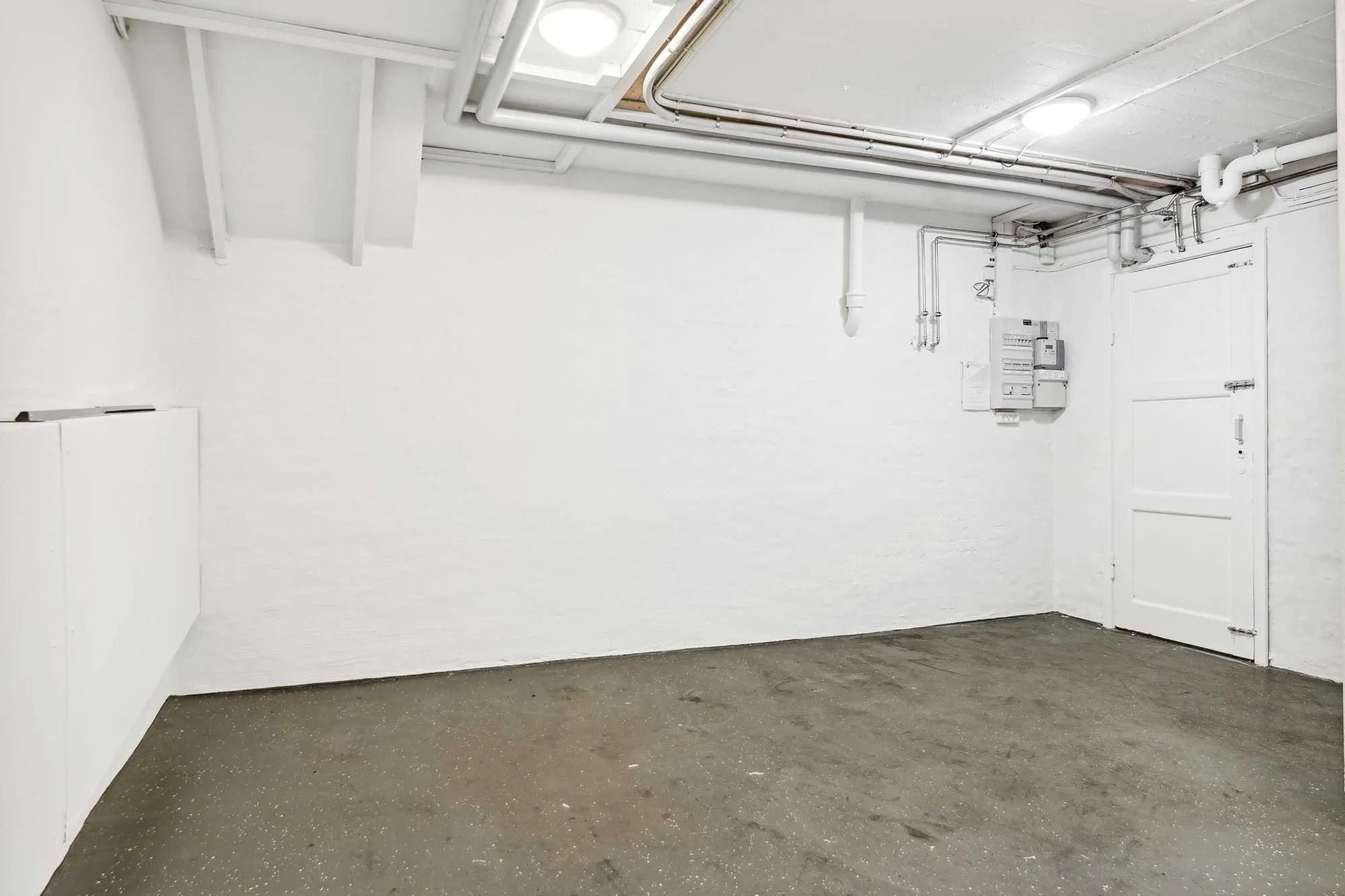 Et tomt rum med hvide vægge og cementgulv samt et lukket hvidt dørblad og flere hvide rør på loftet.