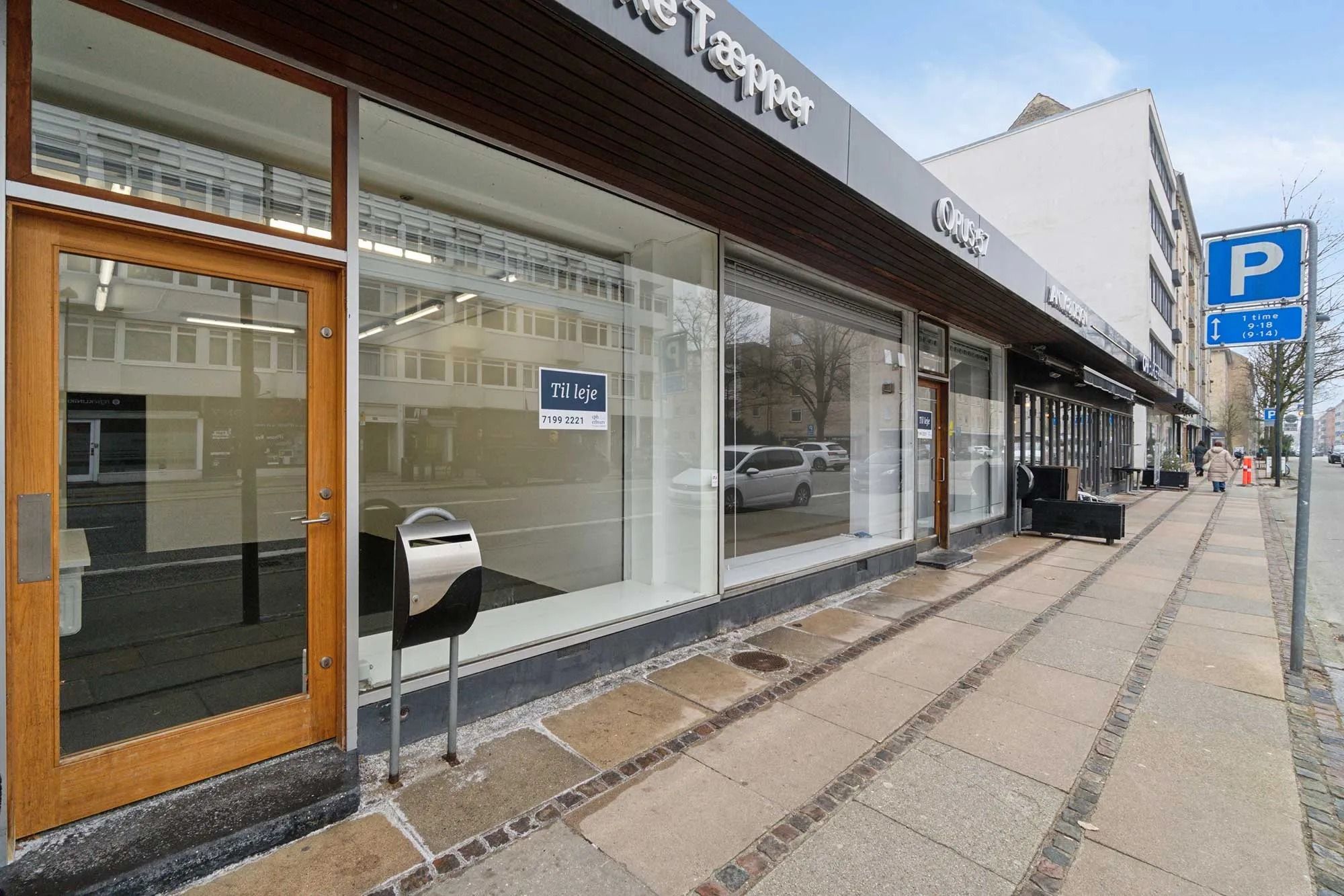 En butik med stort salgsareal og store vinduer fra gulv til loft viser en skilt med "Til leje" og en parkeringsskilt med "P"