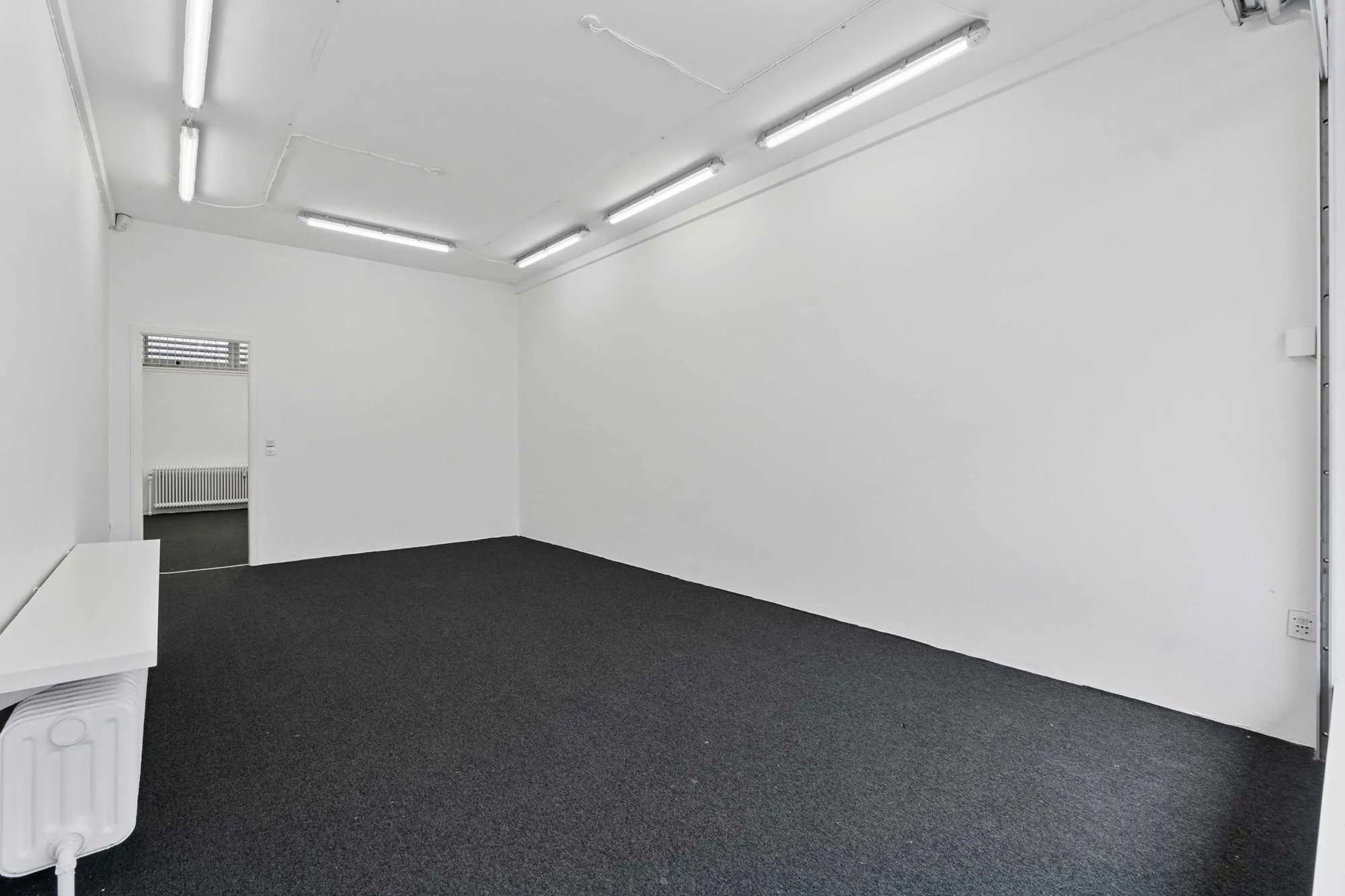 Et lyse, tomt salgsrum med hvide vægge og gulv samt lysekroner i loftet og radiatorer mod væggen