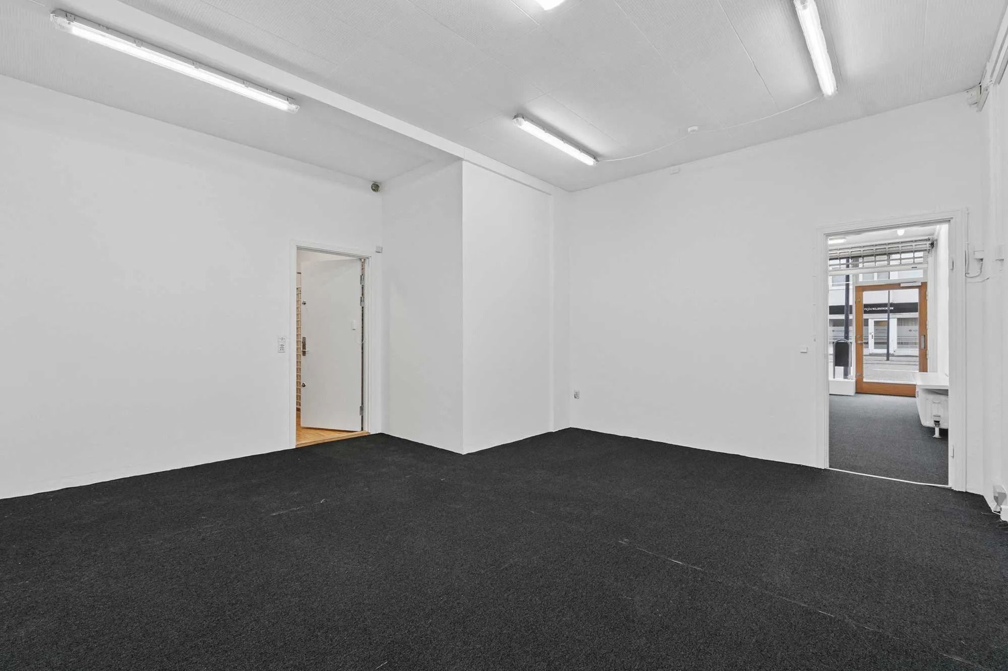 En tom butik med hvide vægge, sort gulv og store vinduer samt lyse, hvide loftslampe og et åbent dørblad til en sidegade