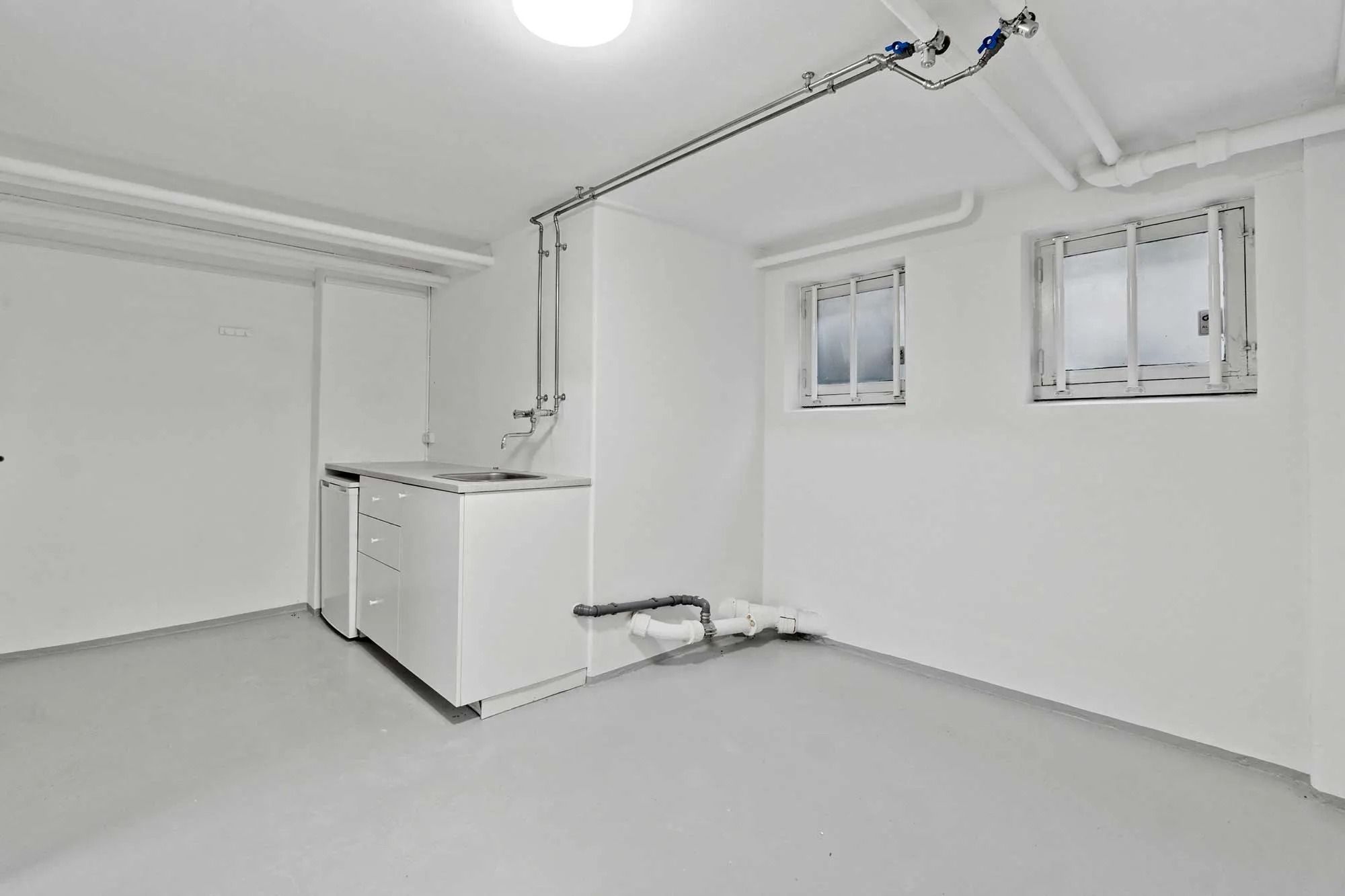 Rummeligt badeværelse med lyse vindue og hvide vægge samt en stor lyskilde og piper og rør i loftet