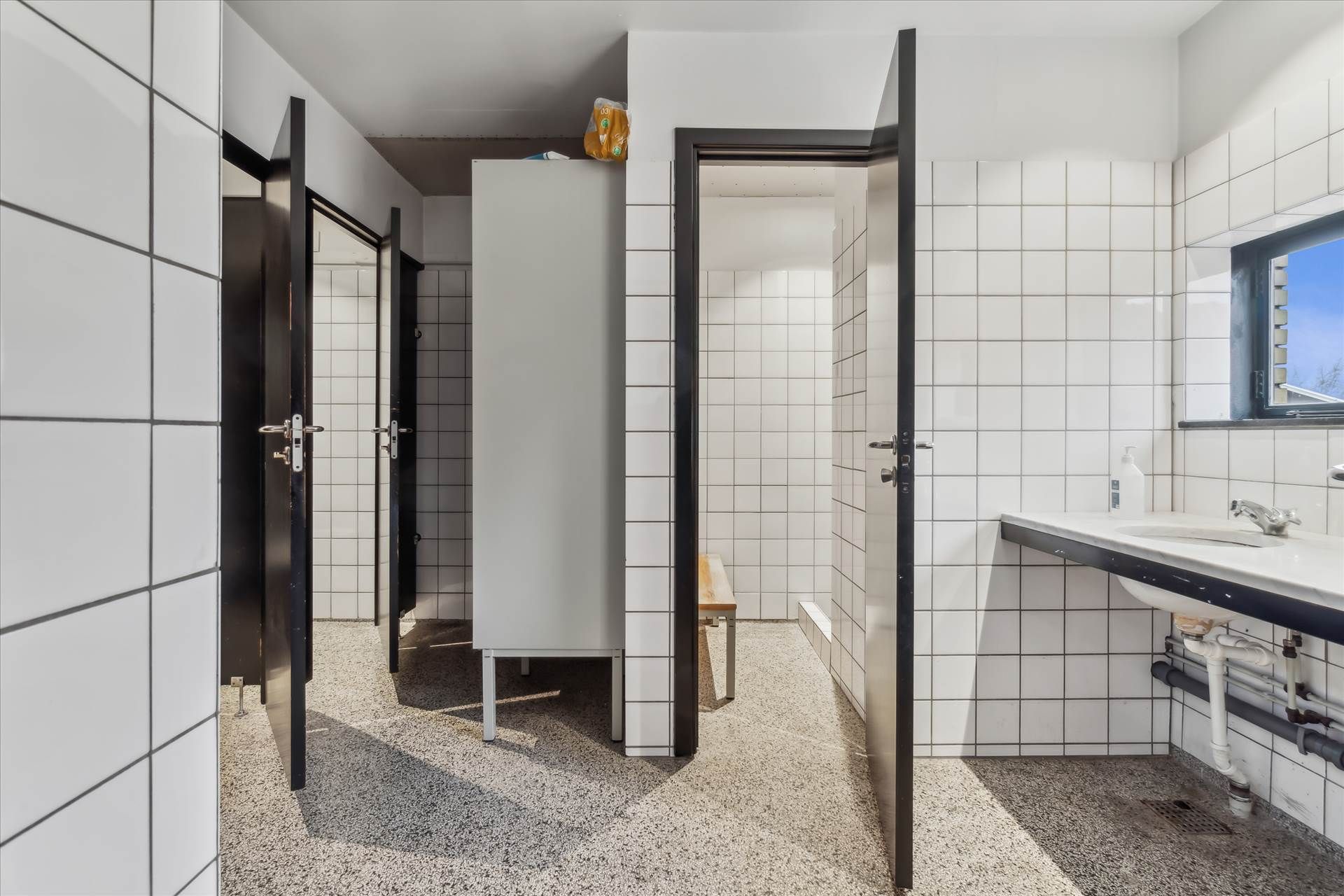 Hvidt og rent toilet med lysegrå gulvbelægning, to åbne døre og en bænk i hjørnet samt et vindue med udsigt til en park