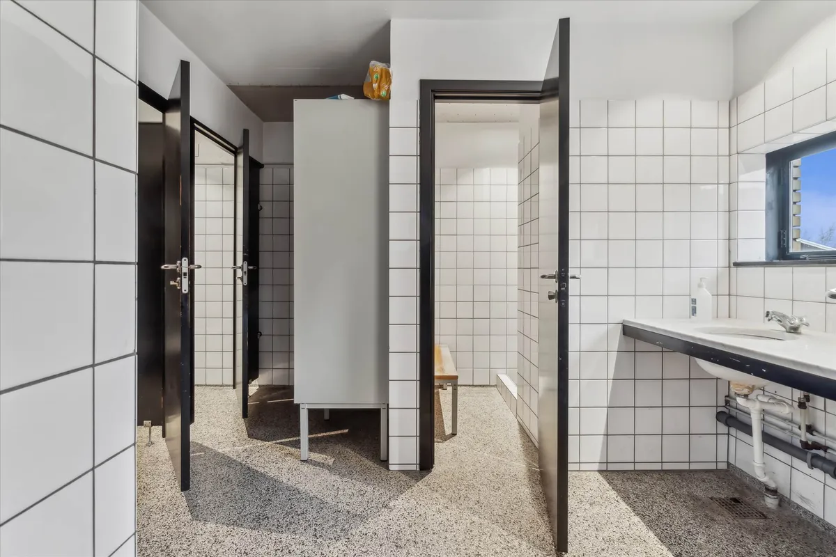 Hvidt og rent toilet med lysegrå gulvbelægning, to åbne døre og en bænk i hjørnet samt et vindue med udsigt til en park