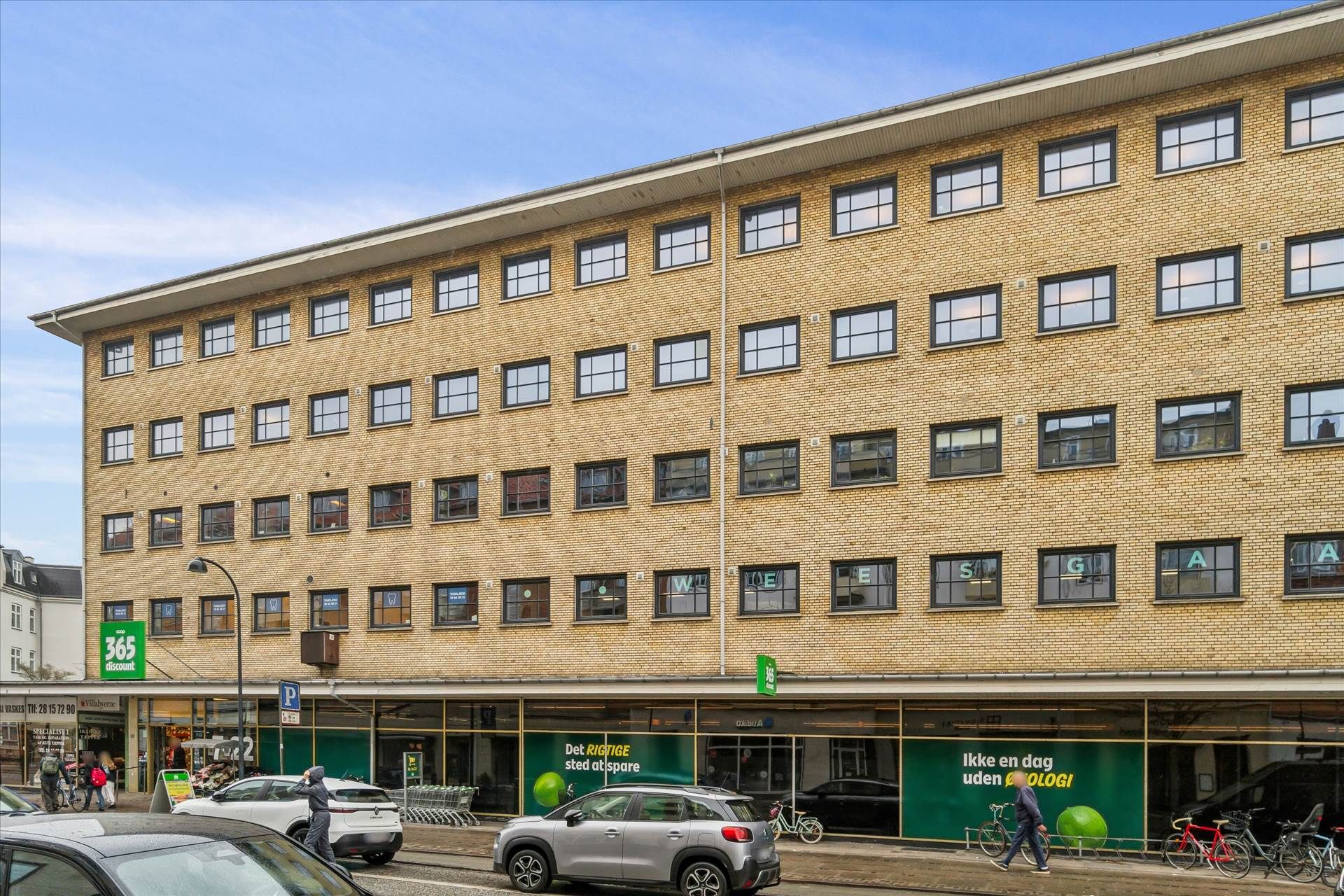 Bygning med flere lejede kontorer og butikker med skilte, biler parkeret udenfor og mennesker og cykler på fortovet