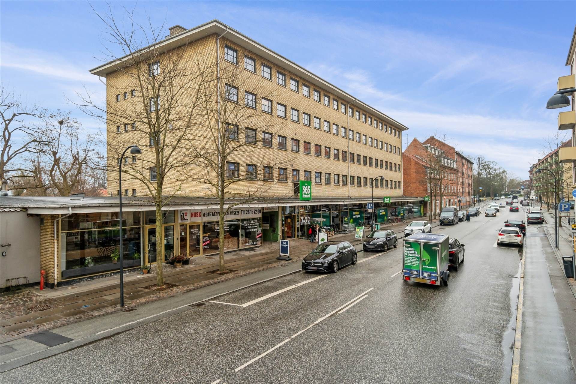 Biler kører på en vej med hvide og gule striber, med grøn skilt og bygning i baggrunden, og søjler med grønt lys