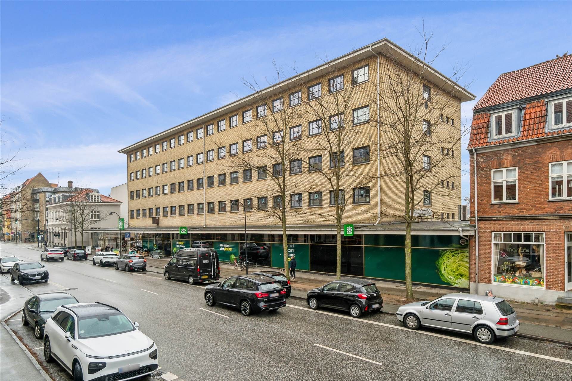 Flere biler parkeret på hver side af en bred gade med butikker og gafvogne på forgrunden