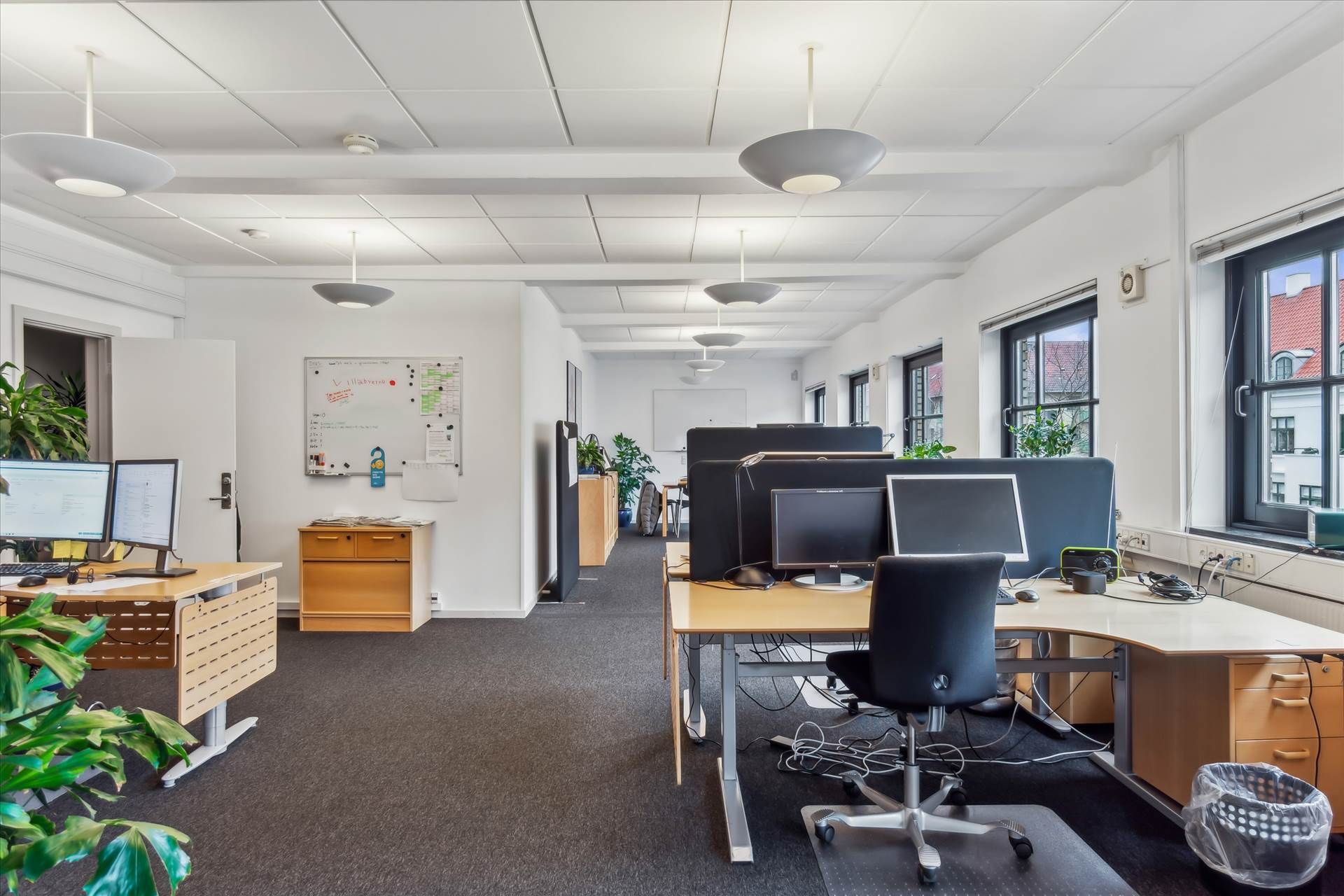 Kontorrum med lyse loftsbelægninger, lysekroner og borde med computere samt flere ruder med udsigt til gaden
