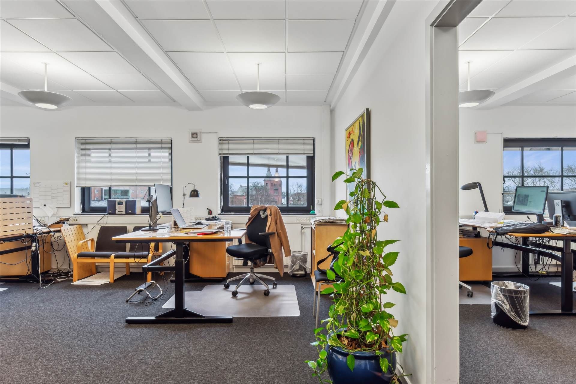 Kontor med bord, stole, computere og plante i et rum med vinduer og lyse gulvbelægning