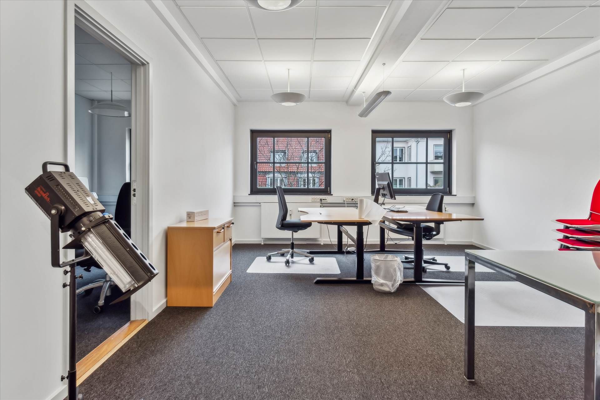 Kontor med to skriveborde og stole samt gulvbelægning, højt hængende lysekroner og vinduer med udsigt til bygninger