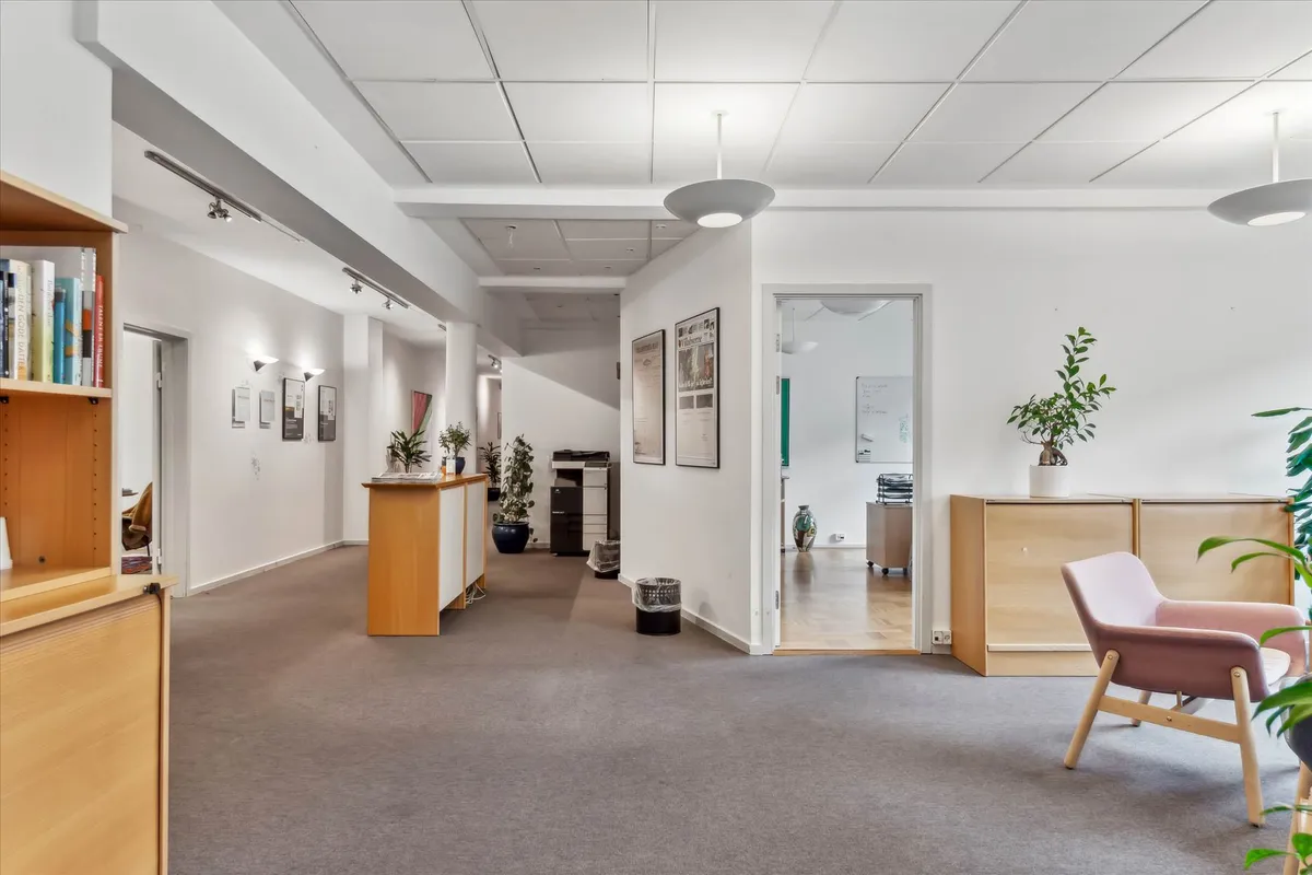 Kontorlokaler med lyse vægge og gulvtæppe samt møbler, der inkluderer et skrivebord, en stol, en sofa, og et skab med plante