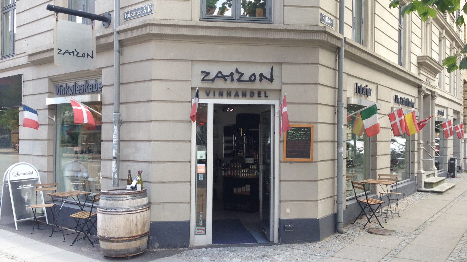 Vinbar Zaizon med stolpe og træfat udenfor og flere vinduer og flag med forskellige lande på facaden