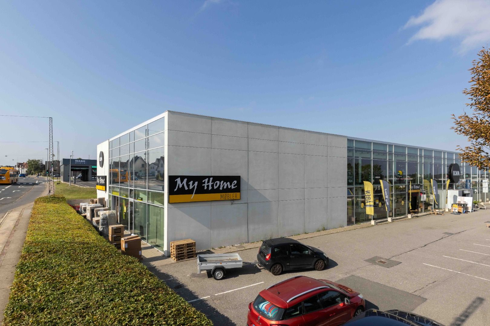 Lys butikslejemål med store vinduer og My Home-skilt, to biler parkeret, træpaletter og guldbrun bus på baggrunden