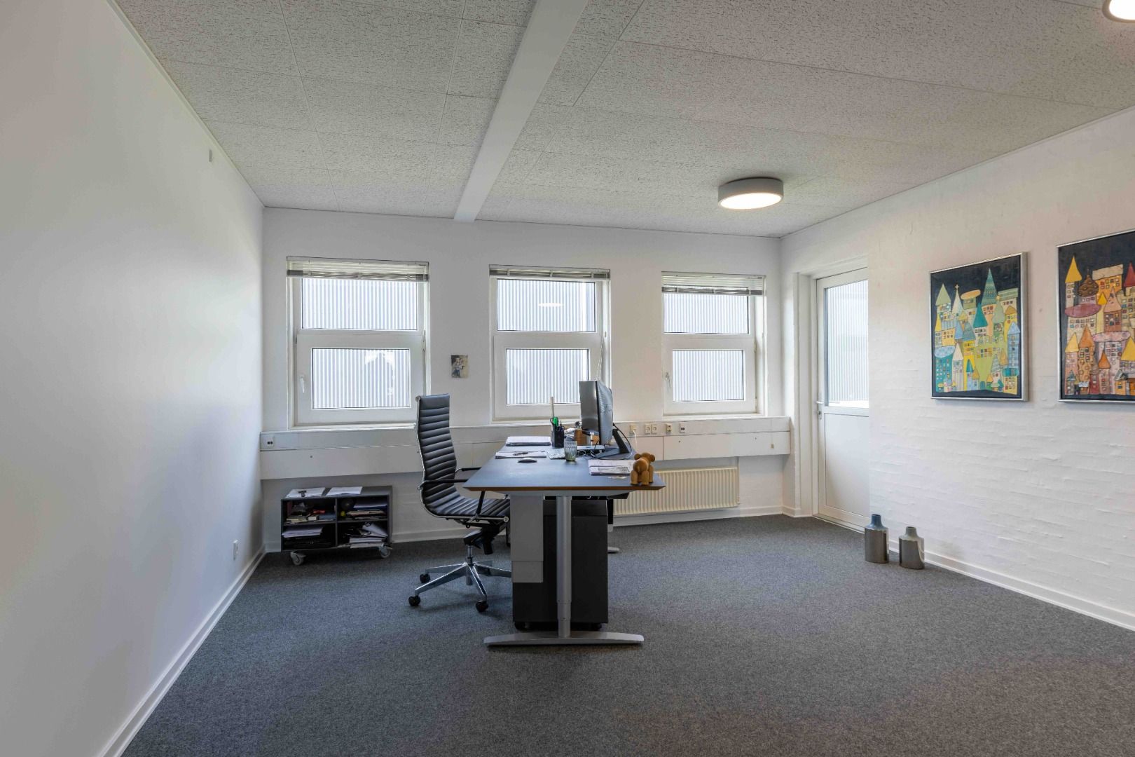 Moderne kontor med to vinduer, skrivebord, stol, trægulv, lysekroner og to malerier.