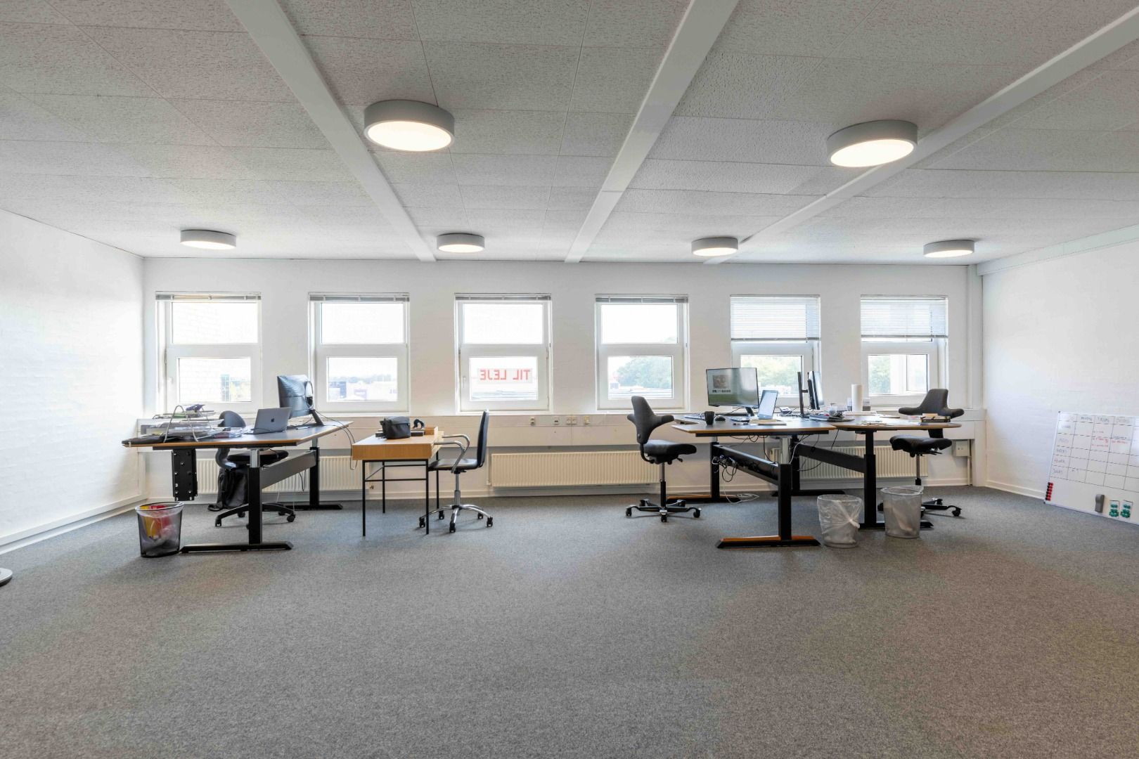 Moderne kontor med tre skriveborde, hver med computere og skrivebordslamper. Væggene er hvide og rummet har lyse loftslampe