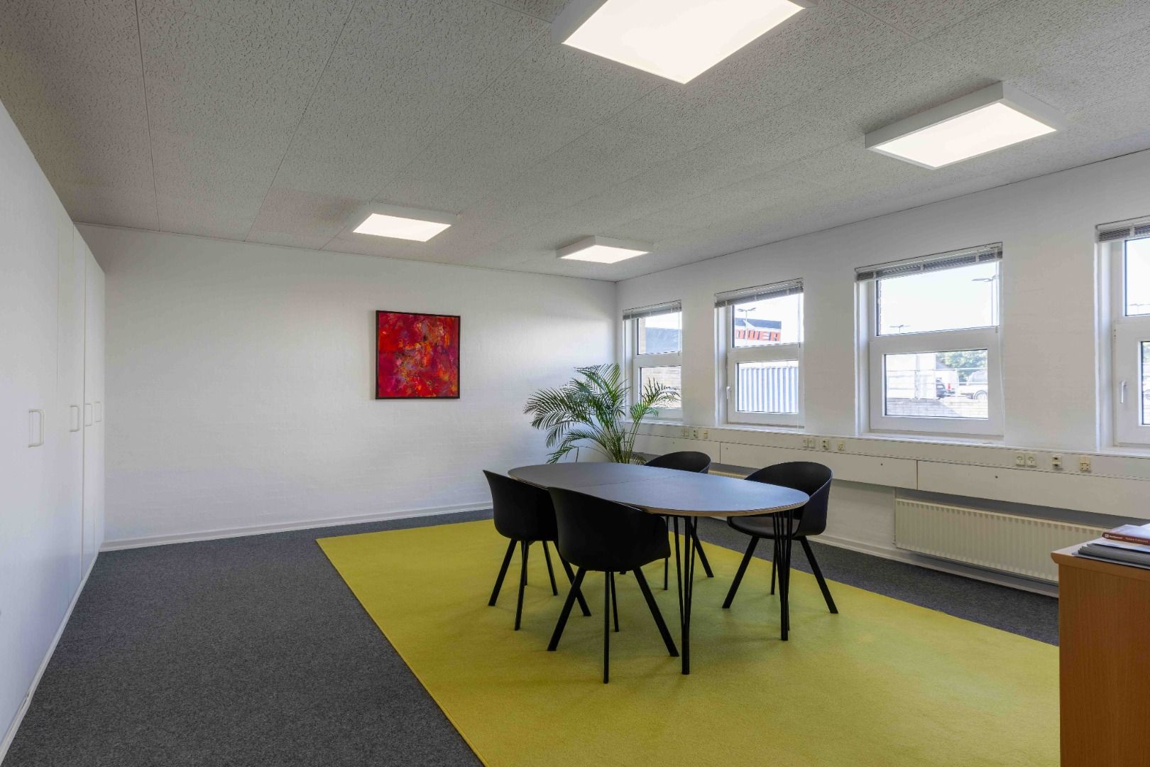 Kontor med rundt bord, fire stole og lysegul gulvtæppe. Væggene er hvide med højloft. Lyse loftslamper, vinduer og plante i
