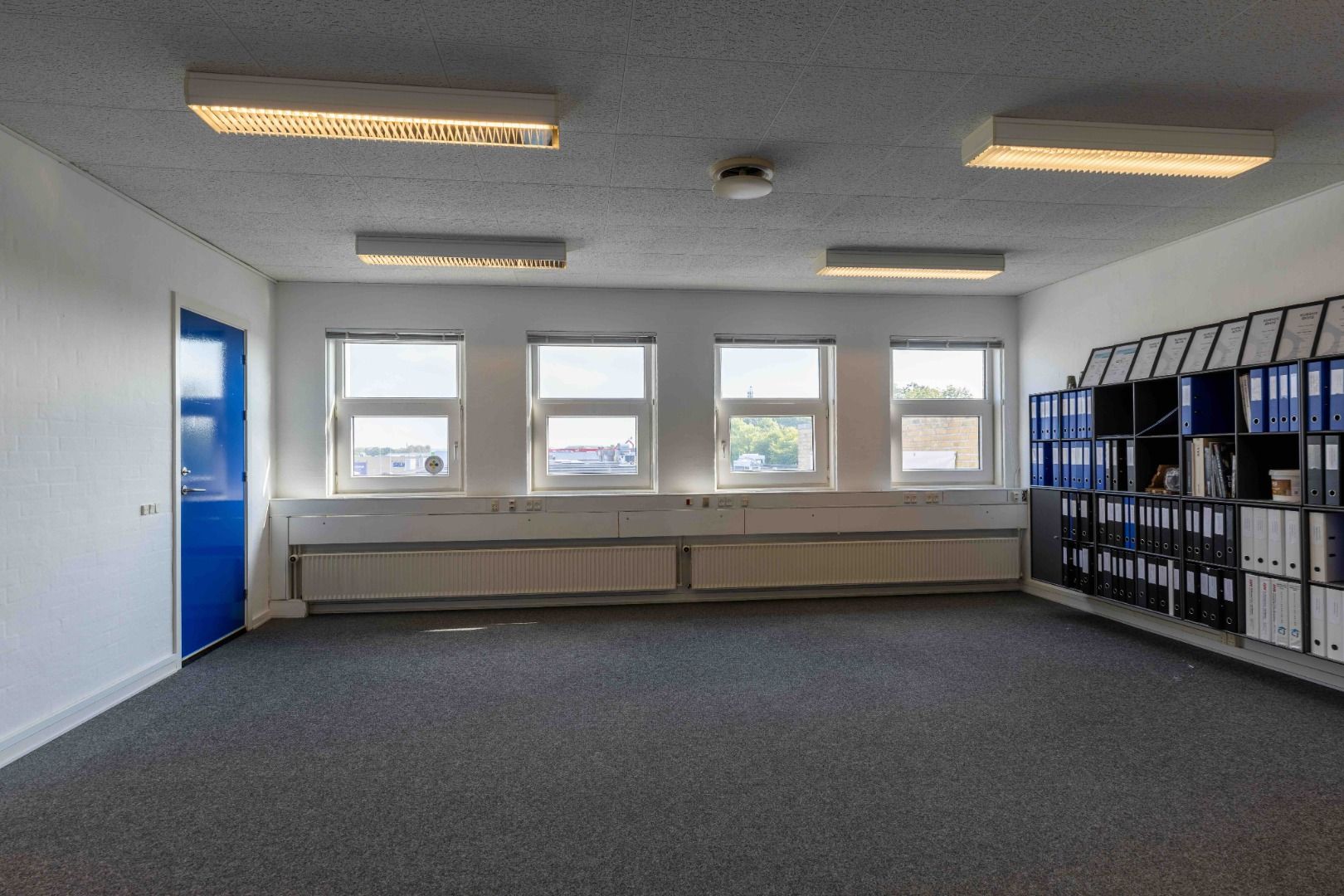 Moderne kontor med tre vinduer, blå dør, hvide vægge, radiatorer og højloftslys. Værelset er tomt med et hyldesystem og