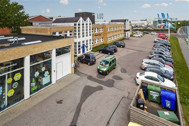 Moderne bygning med glasvinduer og flere parkerede biler og skraldespande. Skilte med tekst og teknologi og vinduer i en