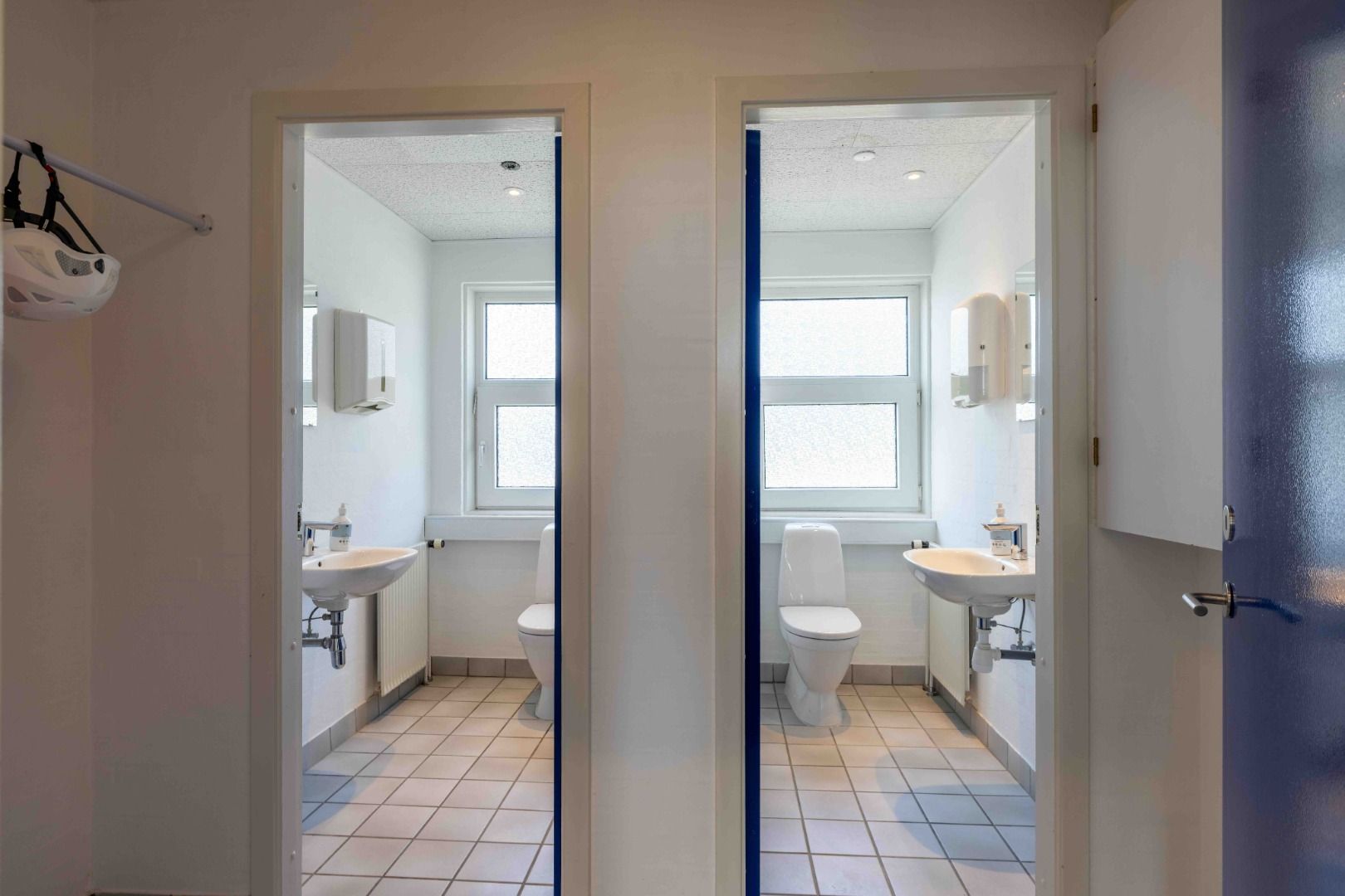 En moderne toiletrum med lyse gulve og hvide vægge, hvor hver toiletrum har en hvid toilet, hængende håndklæder og to hvide