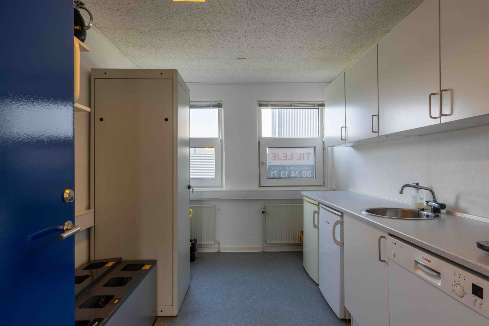 Moderne kontor med køkkendel, hvor der er hvide skabsfronter og højloft. På væggen hænger et skilt med teksten "Cleaning