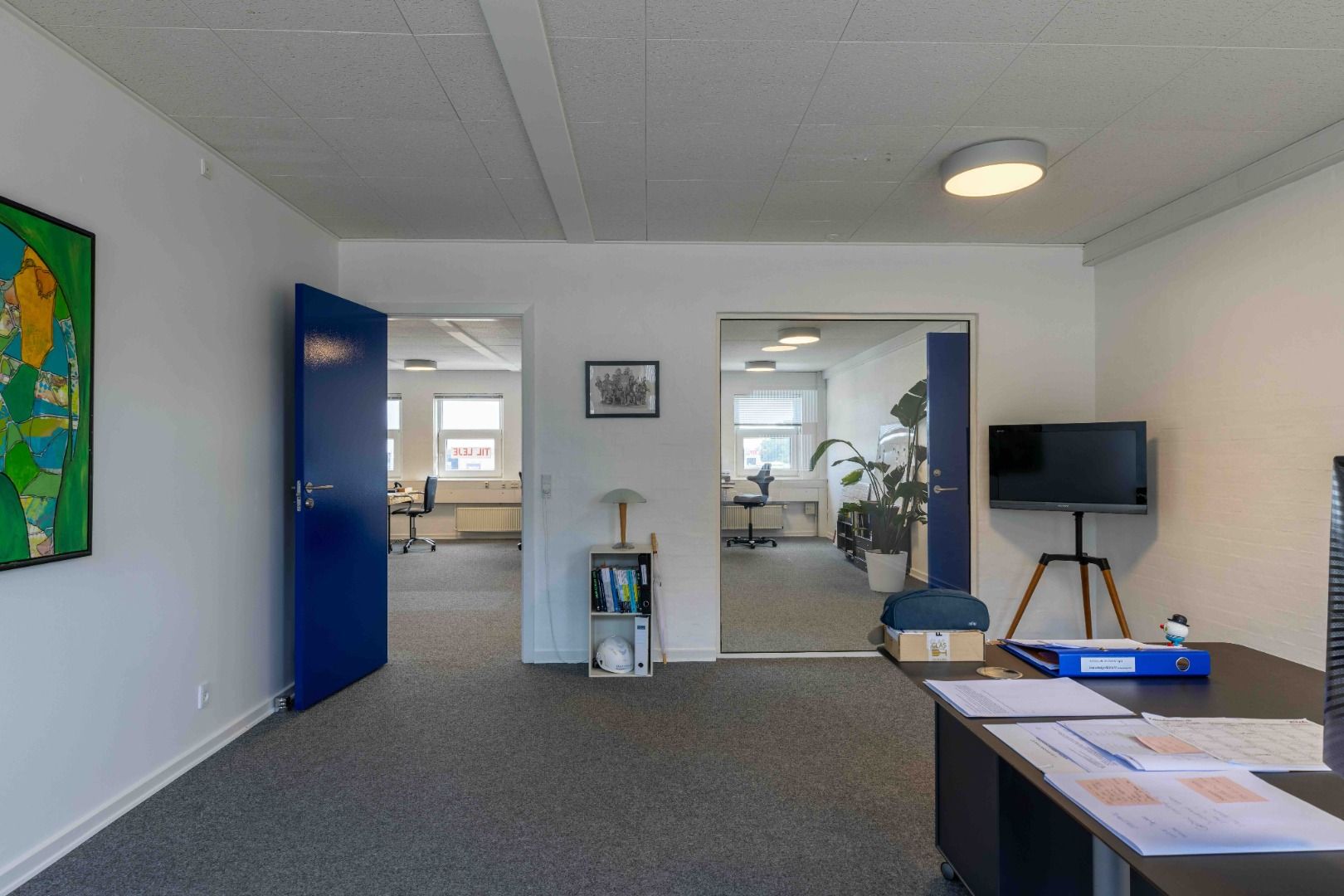 Et moderne kontor med lyse loftslamper og fuldt udstyret skrivebord med papir, mapper og en tv-skærm. En stor maleri og åbne