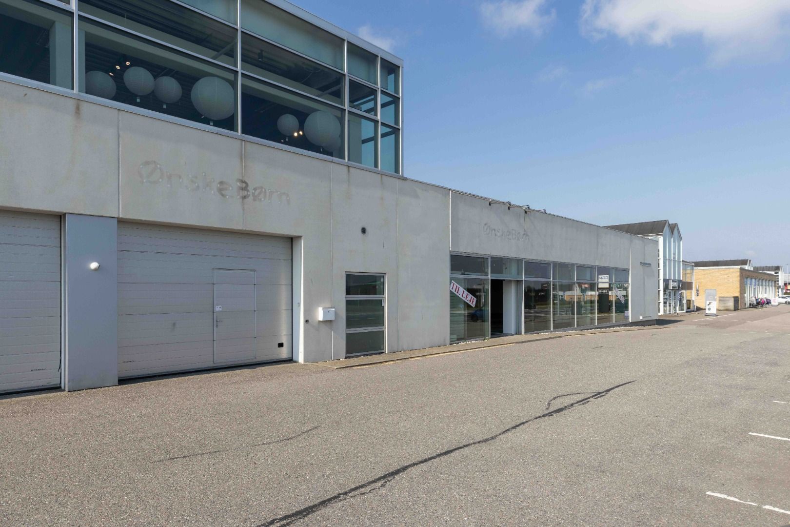 Butikslejemål med to garageporte og flere vinduer, hvor én vinduesdør er åben, og der står "Til Leje" på væggen