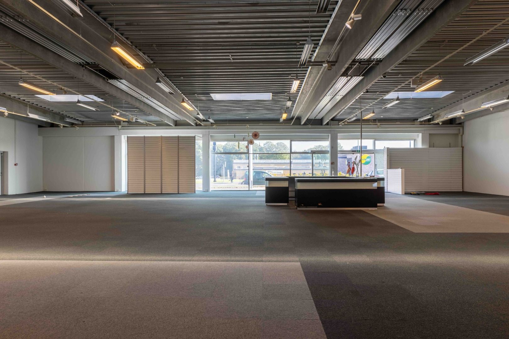 En stor, tom forretningslokal med højloft og glasvinduer, der vender ud mod gaden og bygninger i baggrunden. Gulvet er