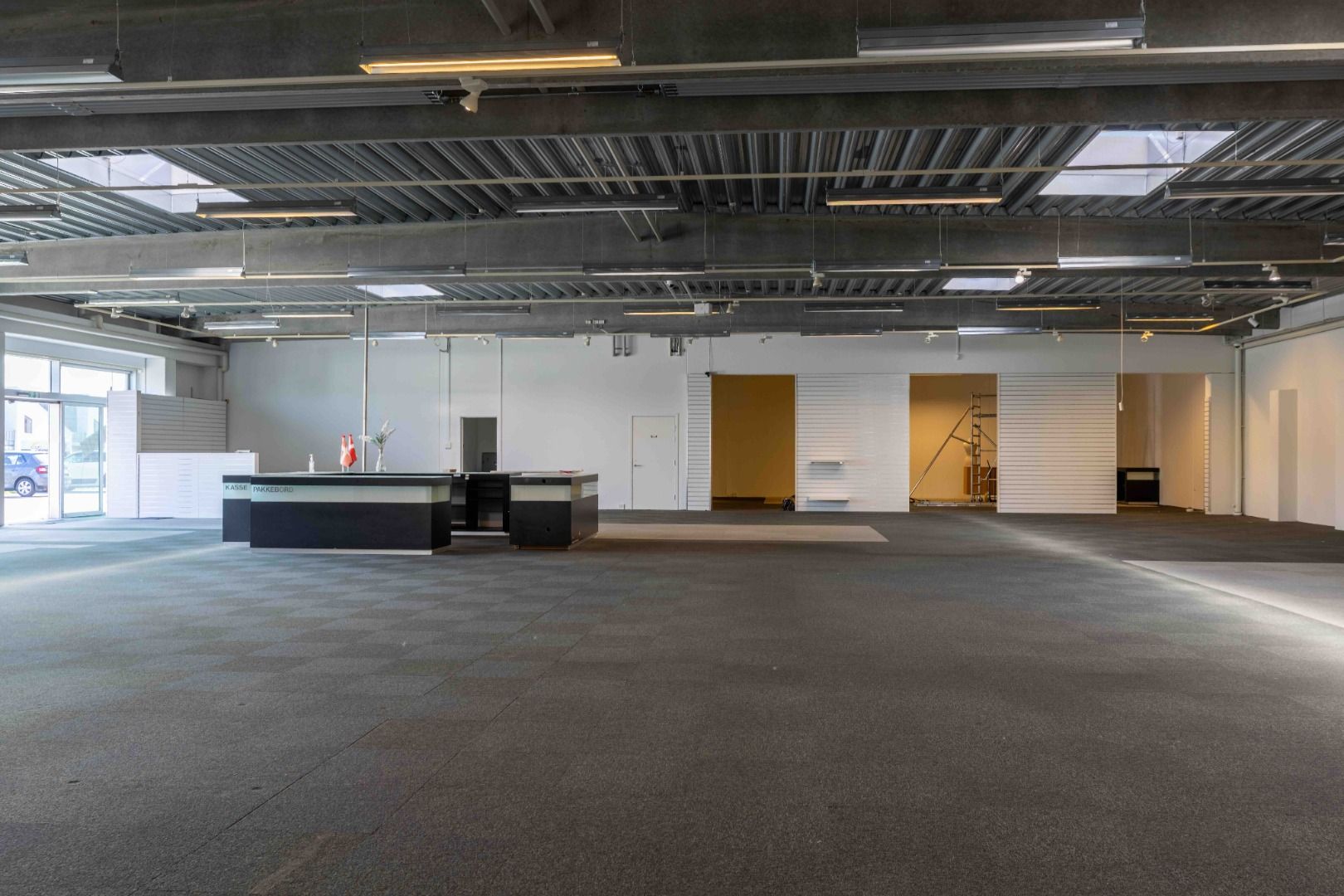 Store kontorlokale med moderne indretning og højt loft. Væggene er hvide, og der er en åbning mod et lagerrum med gul